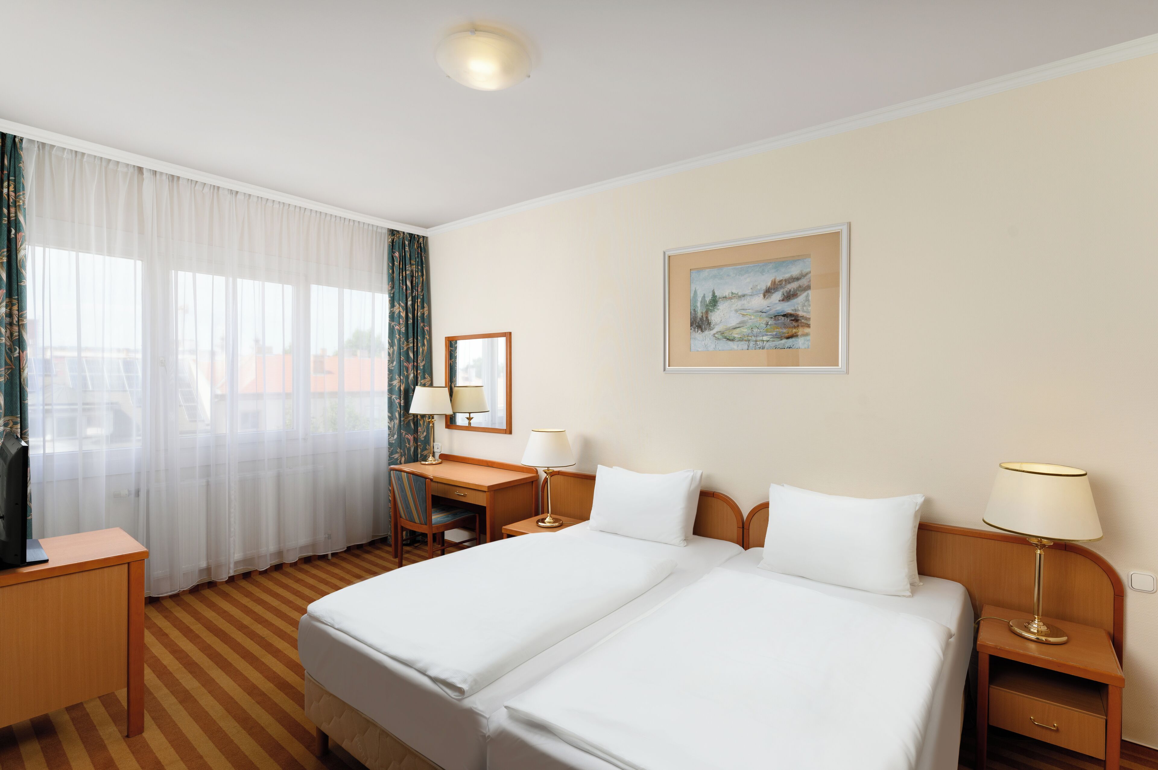 Une chambre d’hôtel avec un grand lit, un bureau, deux tables de chevet avec des lampes, une chaise, une fenêtre avec des rideaux légers et un tableau encadré au mur.