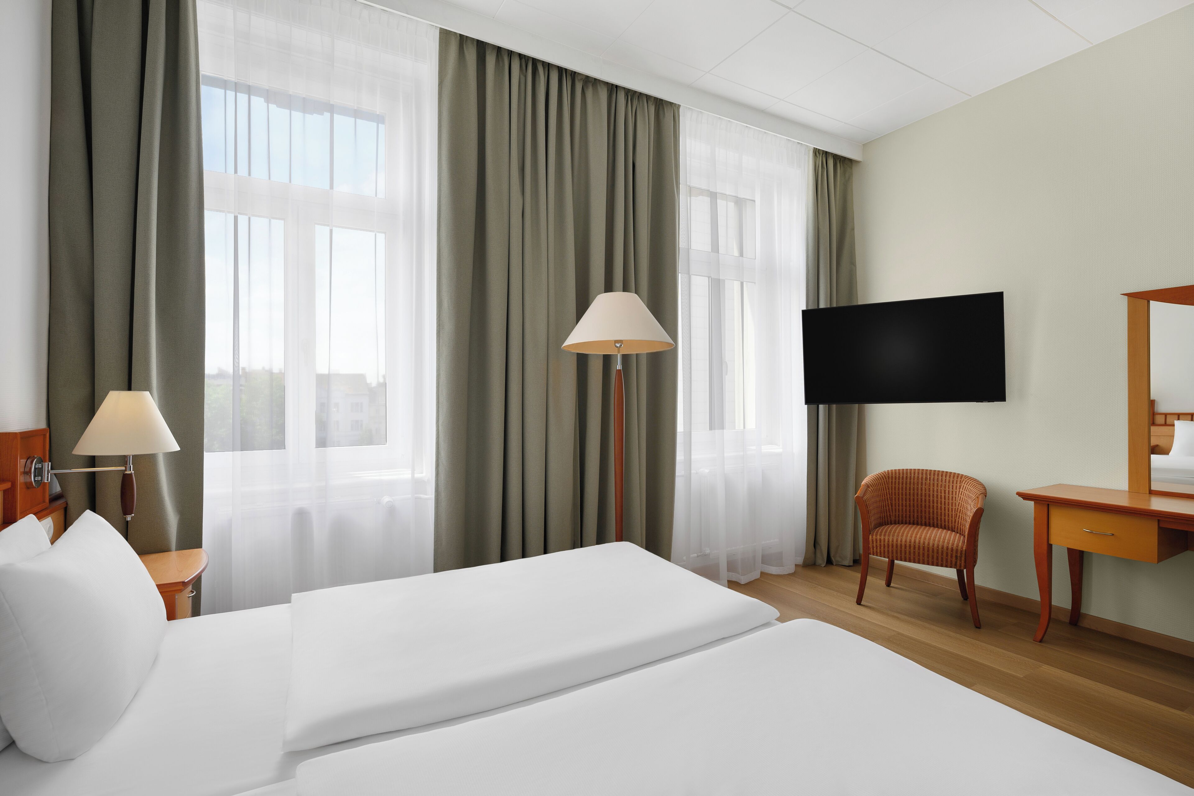 A hotel room with a large bed, a chair, a desk, a TV, and sheer curtains.