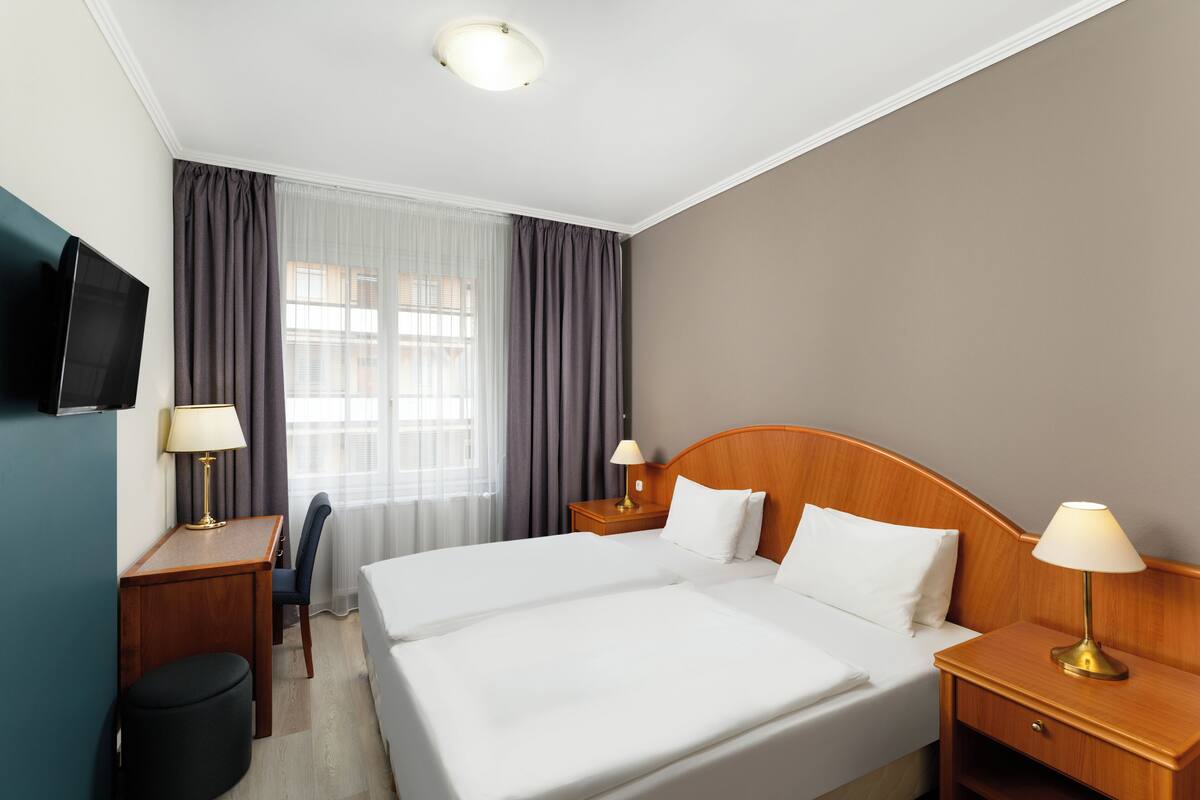 Una habitación de hotel con una cama grande, dos mesitas de noche con lámparas, un escritorio con silla, un televisor montado en la pared y una ventana con cortinas.