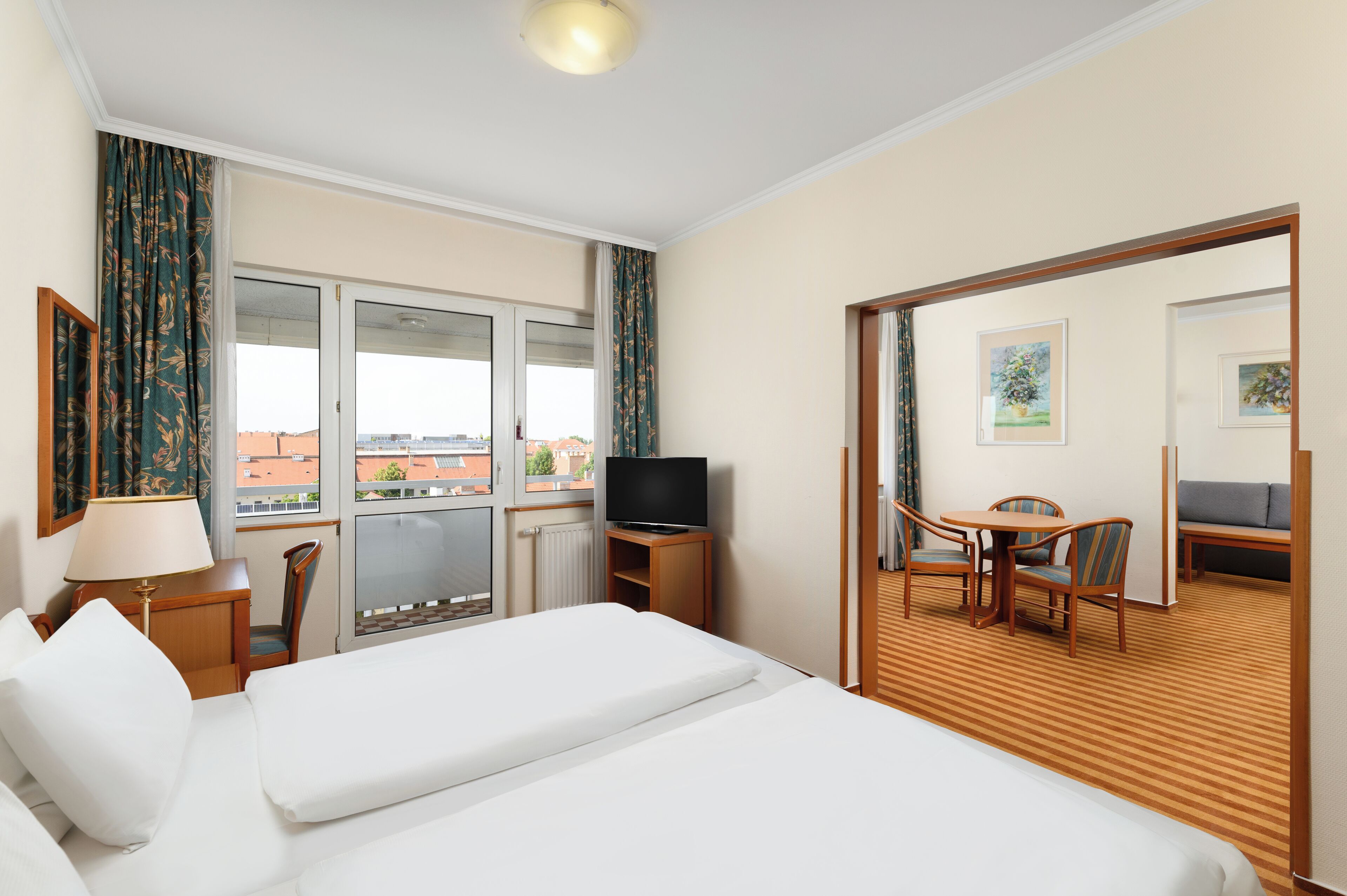 A hotel room with a bed, a TV, a desk with a chair, and a view of the city through large windows.