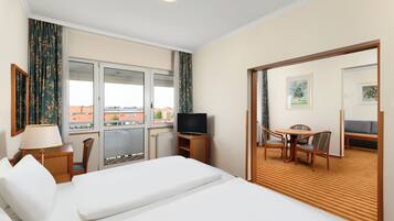 A hotel room with a bed, a TV, a desk with a chair, and a view of the city through large windows.