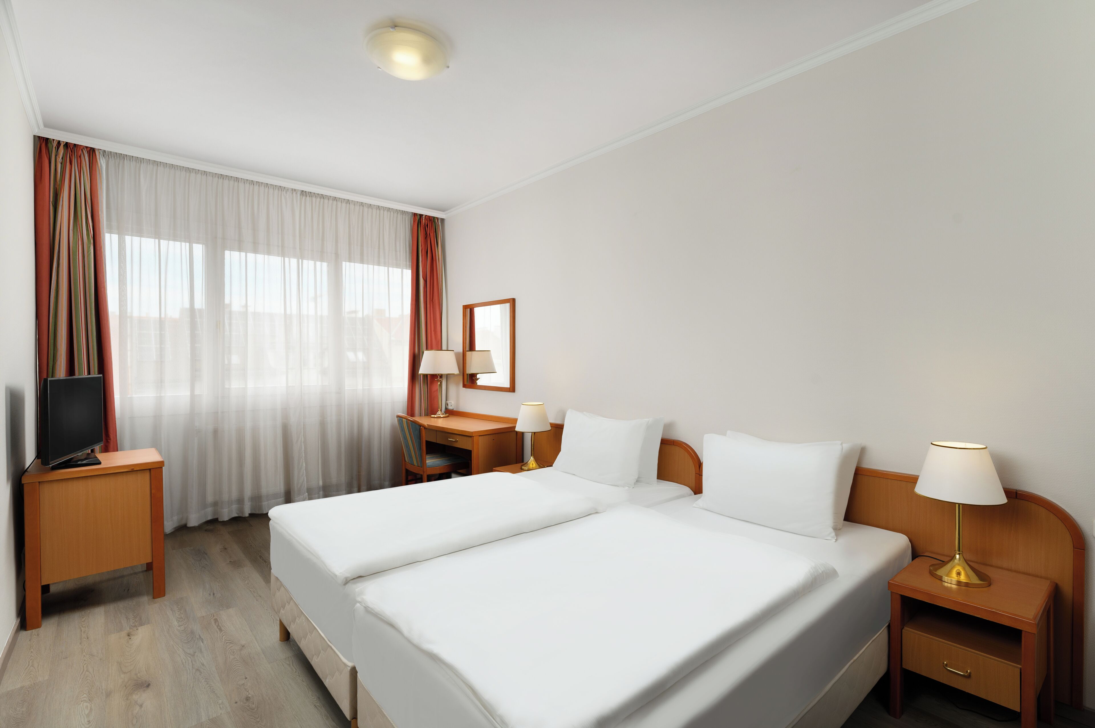 A hotel room with a double bed, a desk with a lamp, a TV, and a large window with curtains.