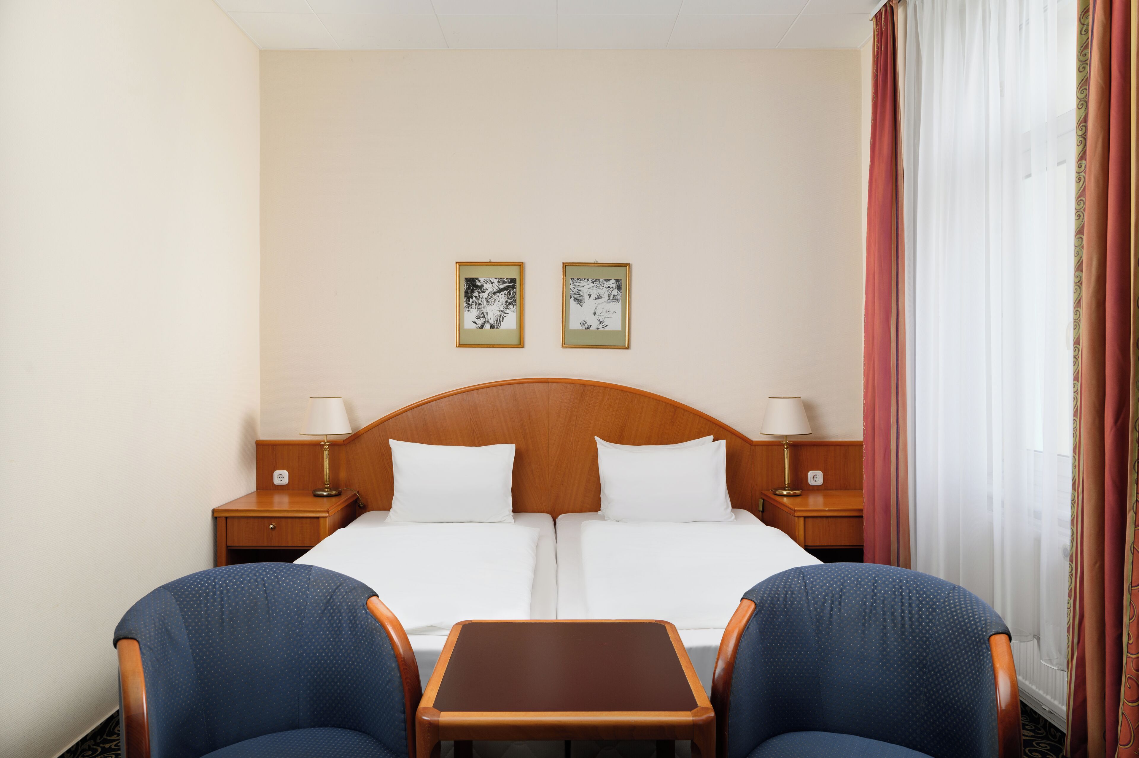 A hotel room with a double bed, two nightstands, a small table, two chairs, and two framed pictures on the wall.