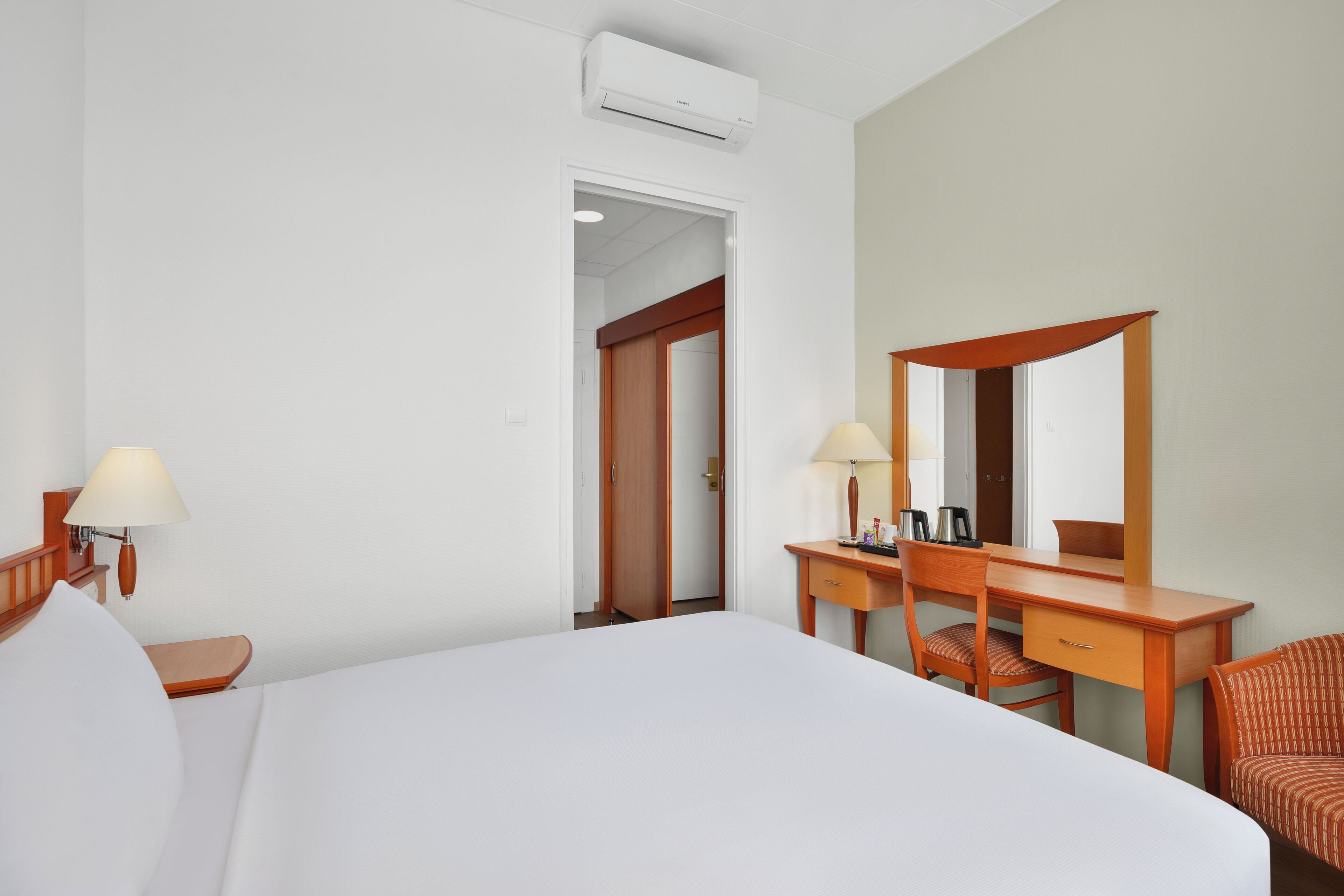 A hotel room with a large bed, a wooden desk, a chair, a mirror, and a lamp.