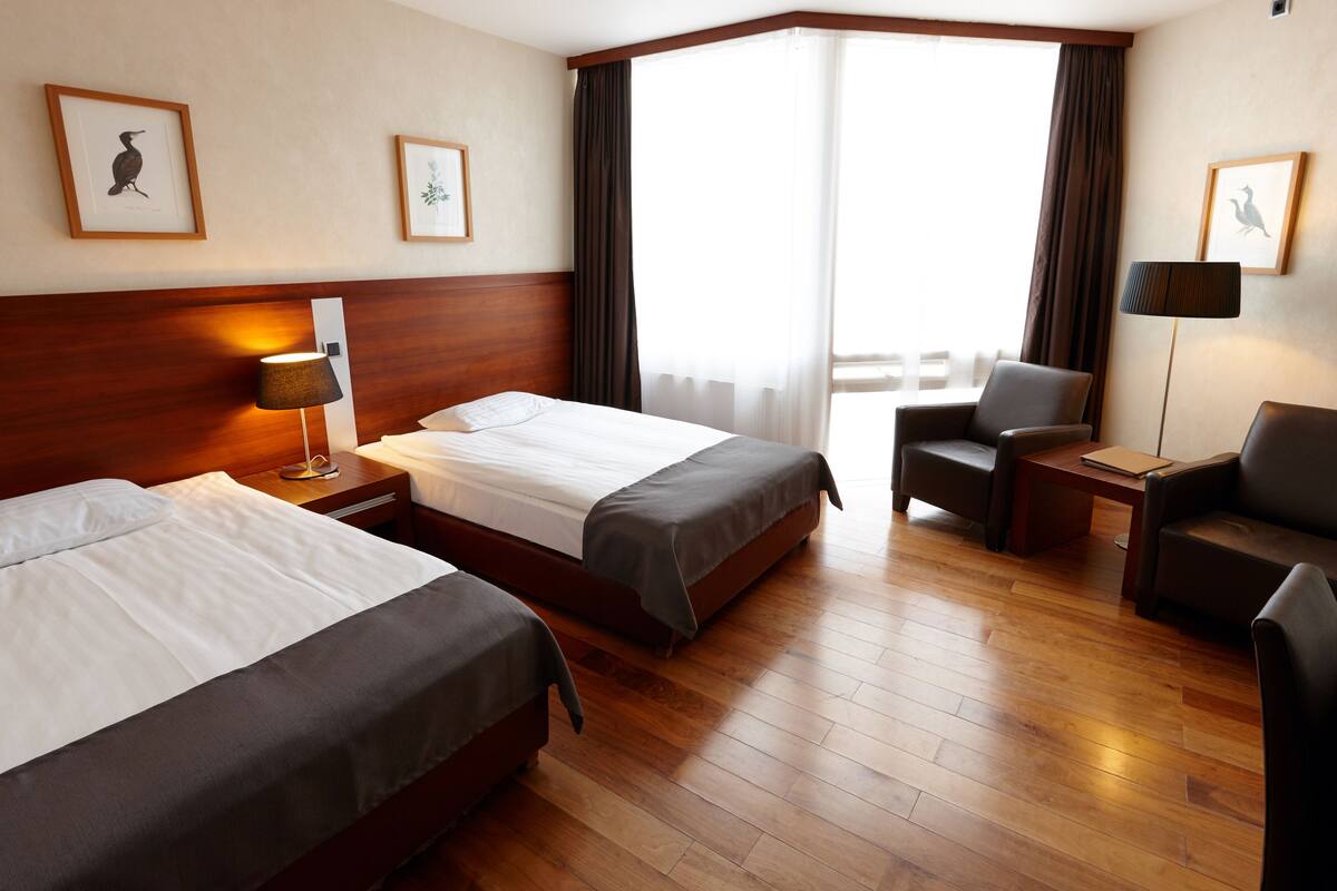 A hotel room with two beds, a desk, a chair, and a large window with curtains.