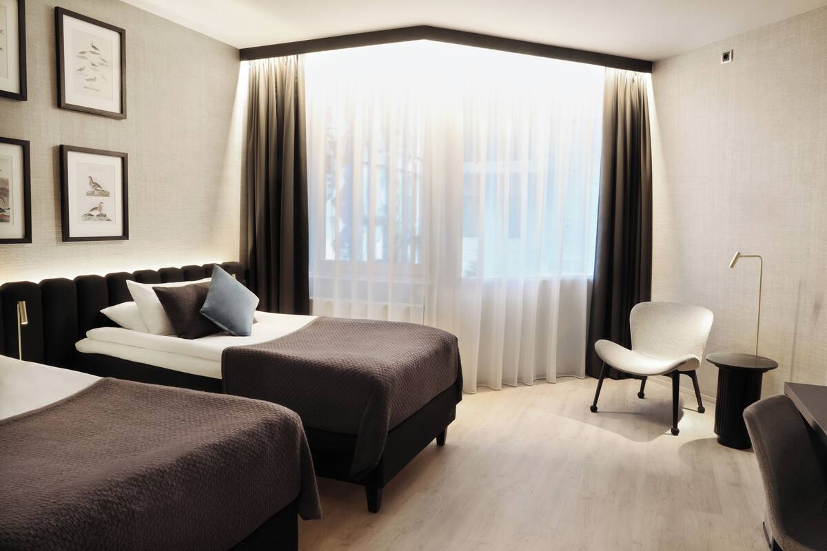 Una habitación de hotel con dos camas, una silla, una mesita y una ventana con cortinas.