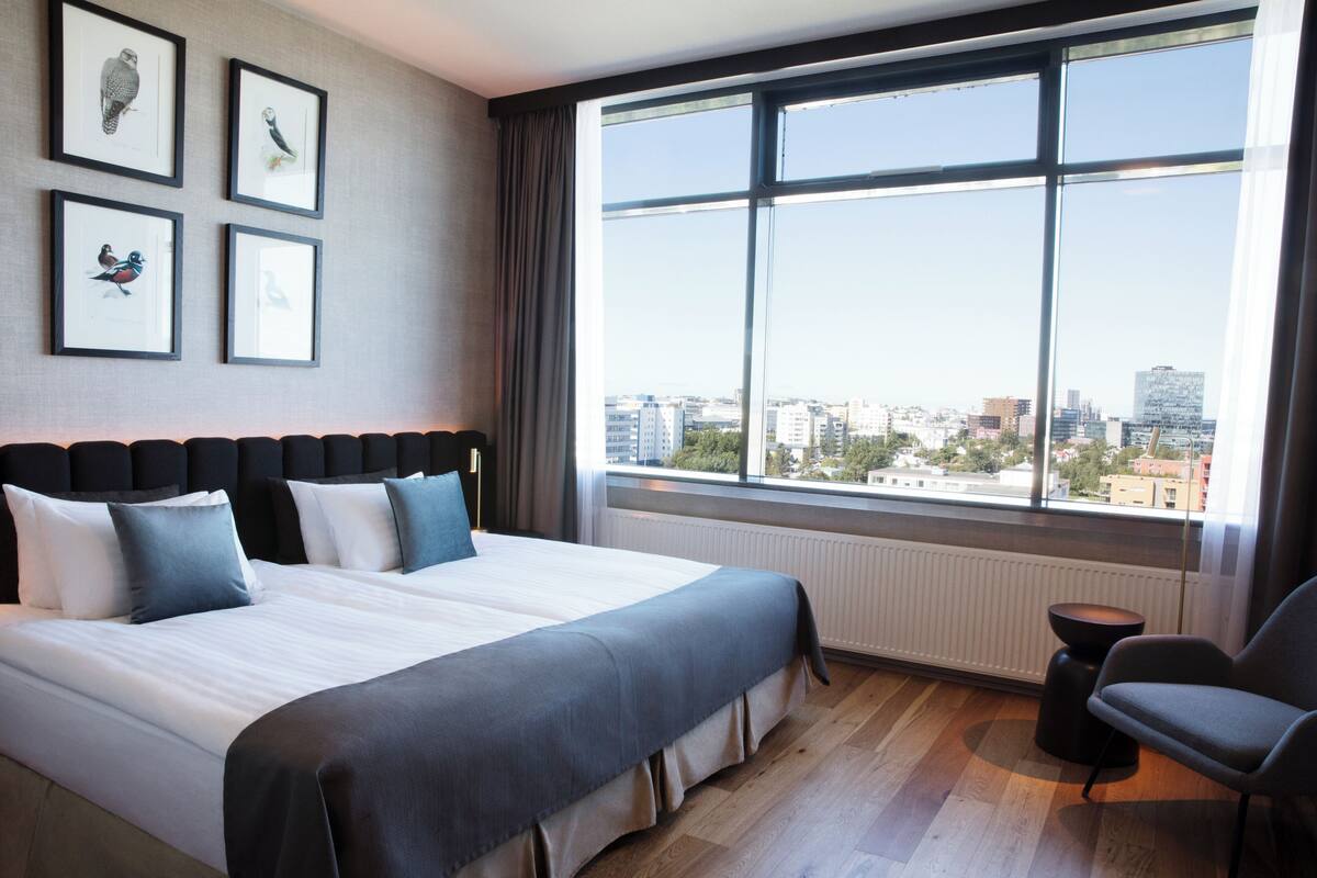 A hotel room with a large bed, a view of the city through a large window, and framed pictures on the wall.