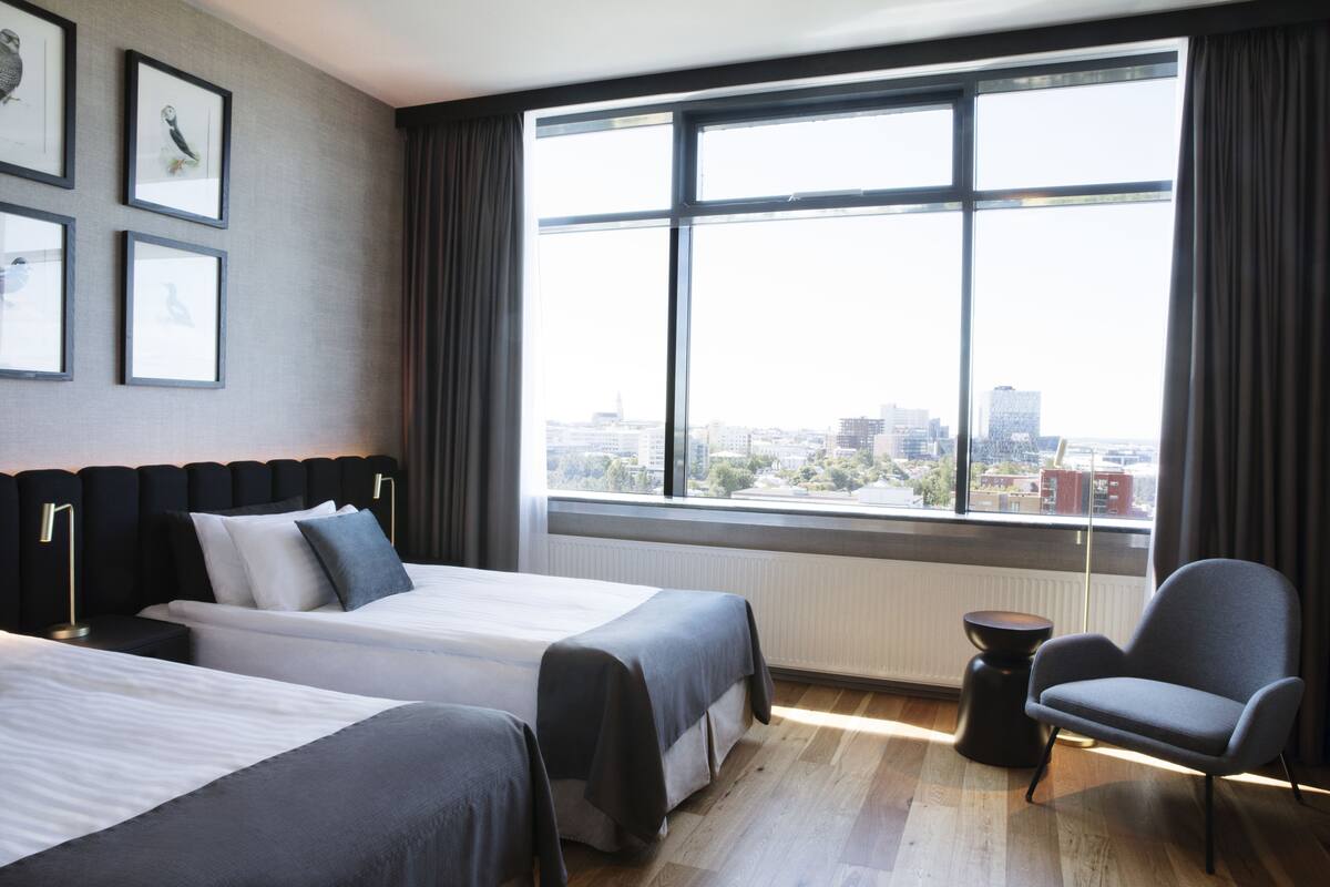 A hotel room with two beds, a chair, a small table, and a large window with a city view.