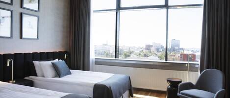 A hotel room with two beds, a chair, a small table, and a large window with a city view.