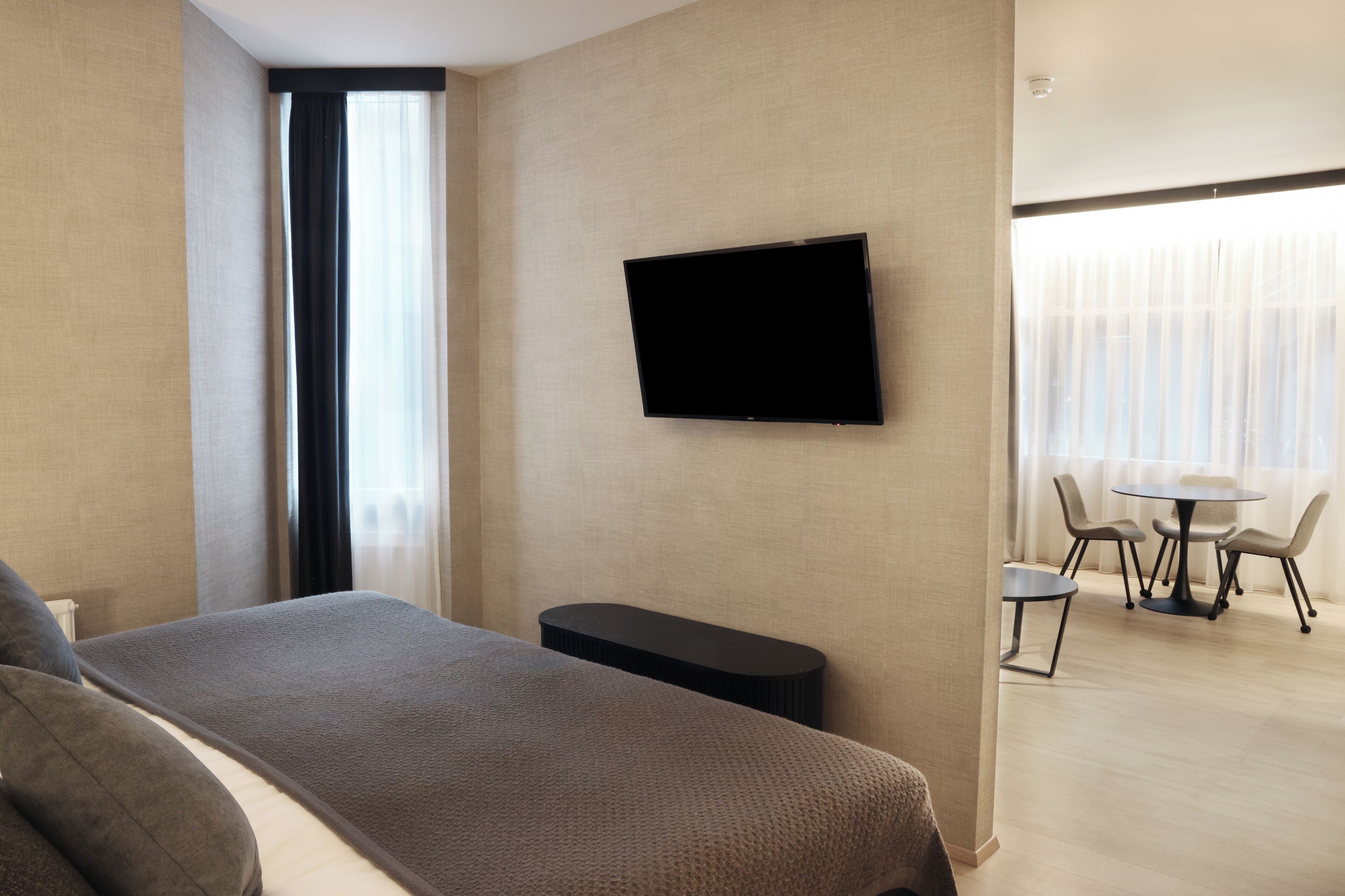 A hotel room with a bed, a TV mounted on the wall, a small table with chairs, and a view of the outside through the window.