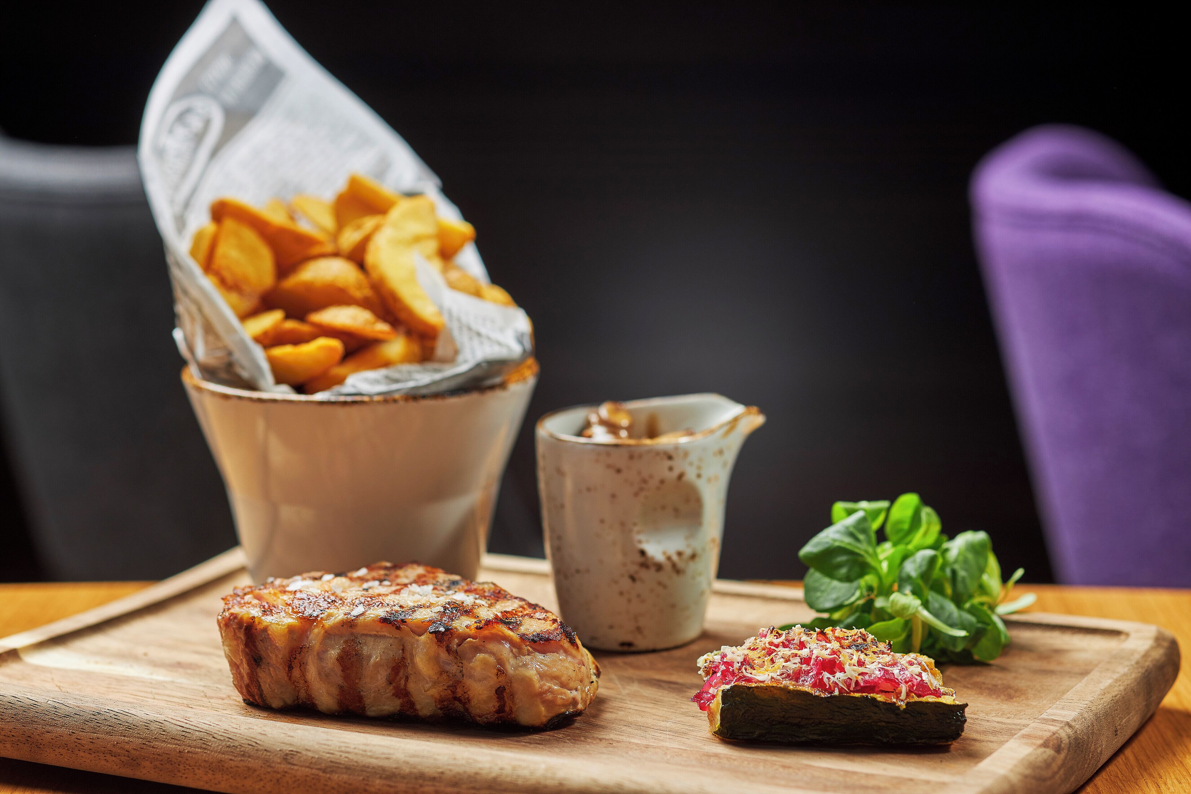 Una tabla de madera con carne a la parrilla, un tazón de papas fritas, una taza pequeña de salsa y algunas verduras sobre una mesa.