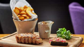 Una tabla de madera con carne a la parrilla, un tazón de papas fritas, una taza pequeña de salsa y algunas verduras sobre una mesa.