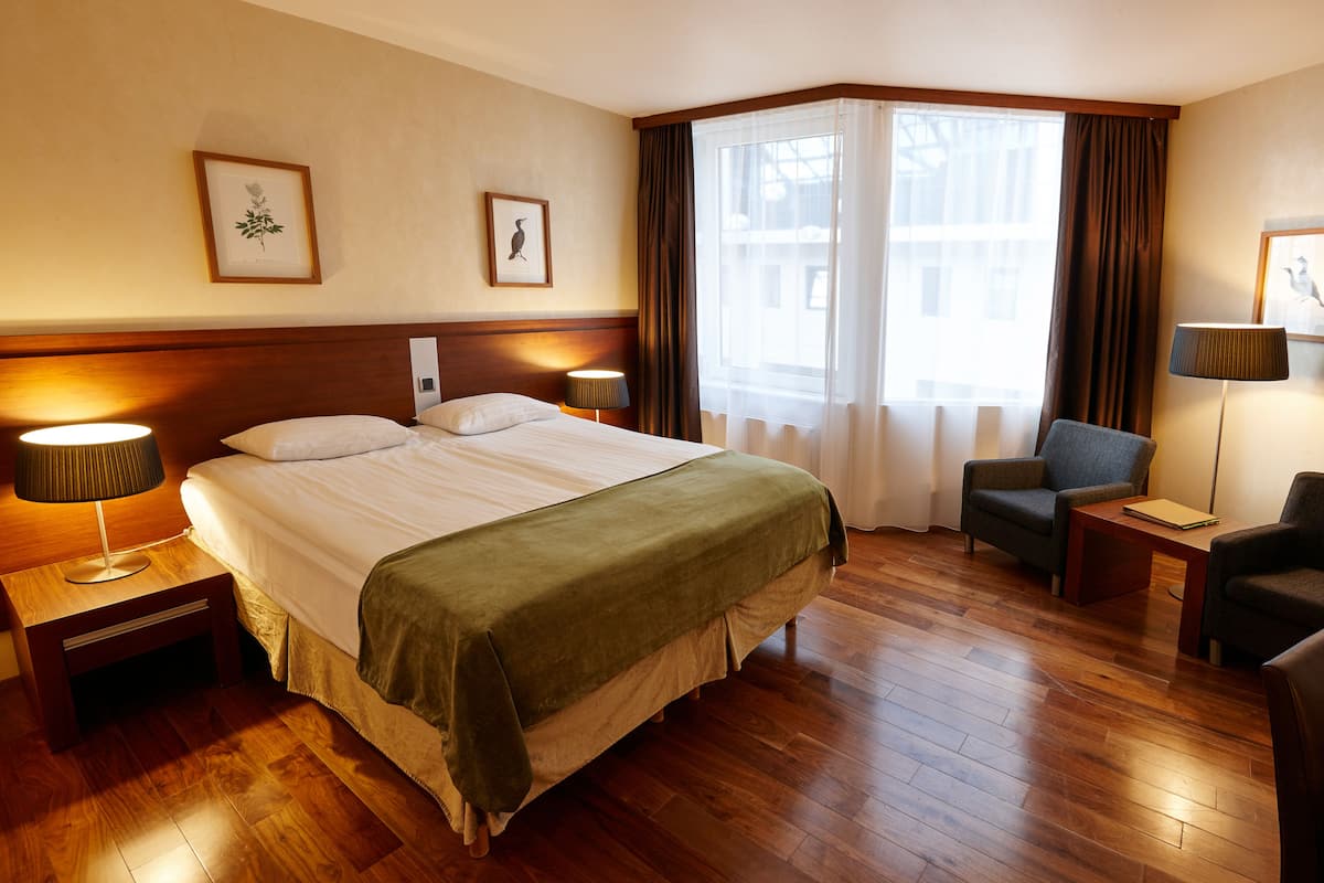 Una habitación de hotel con una cama grande, dos mesitas de noche con lámparas, una silla, una pequeña mesa y una ventana con cortinas.