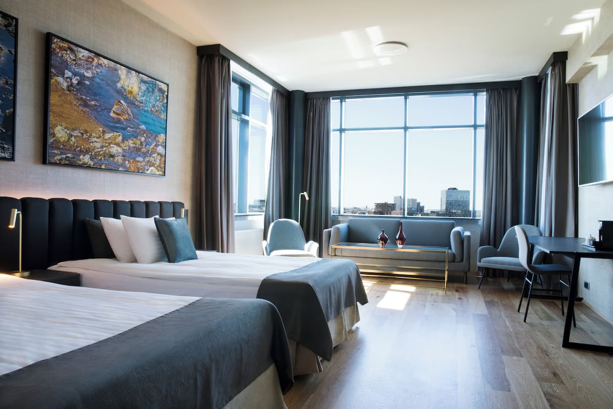 Habitación de hotel con dos camas, un sofá, un escritorio y un amplio ventanal con vista a la ciudad.