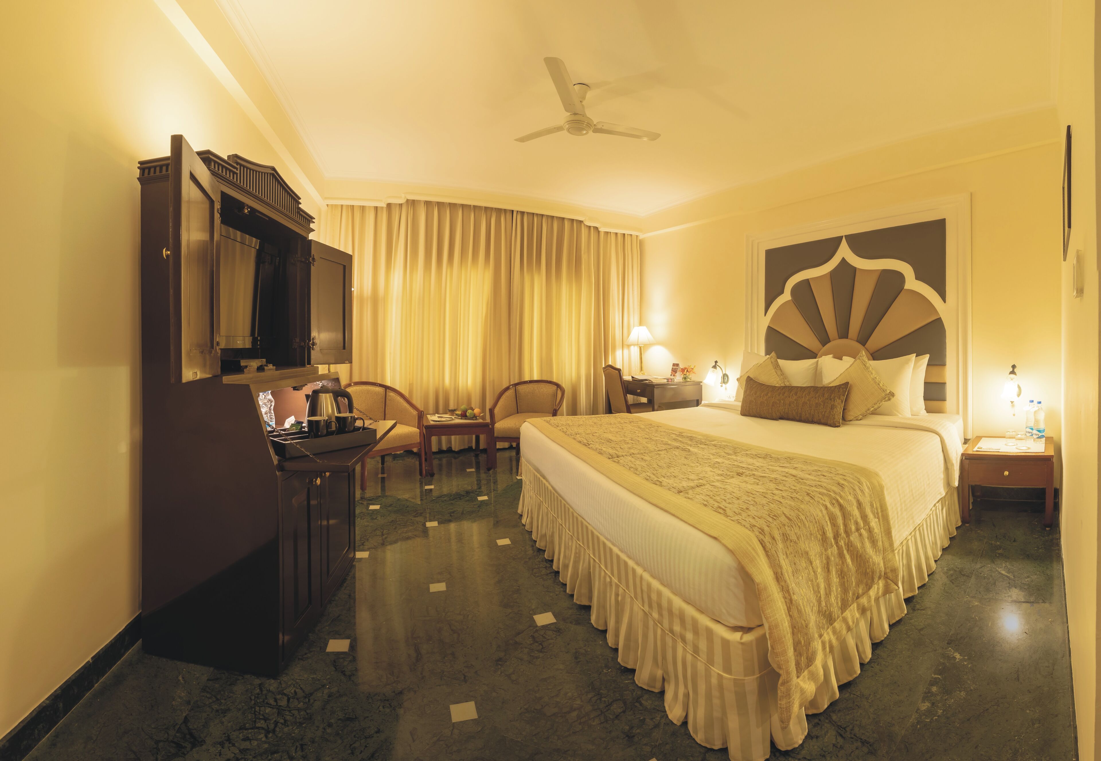A hotel room with a large bed, a desk with a lamp, a chair, and a television.