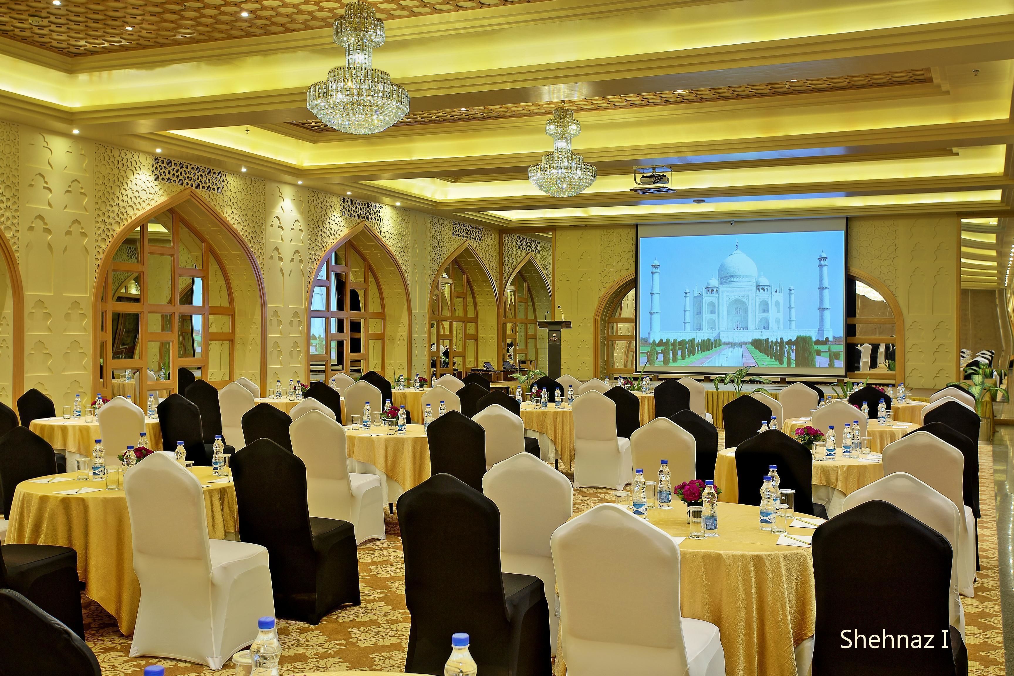A banquet hall with round tables, black and white chairs, chandeliers, and a large screen displaying the Taj Mahal.