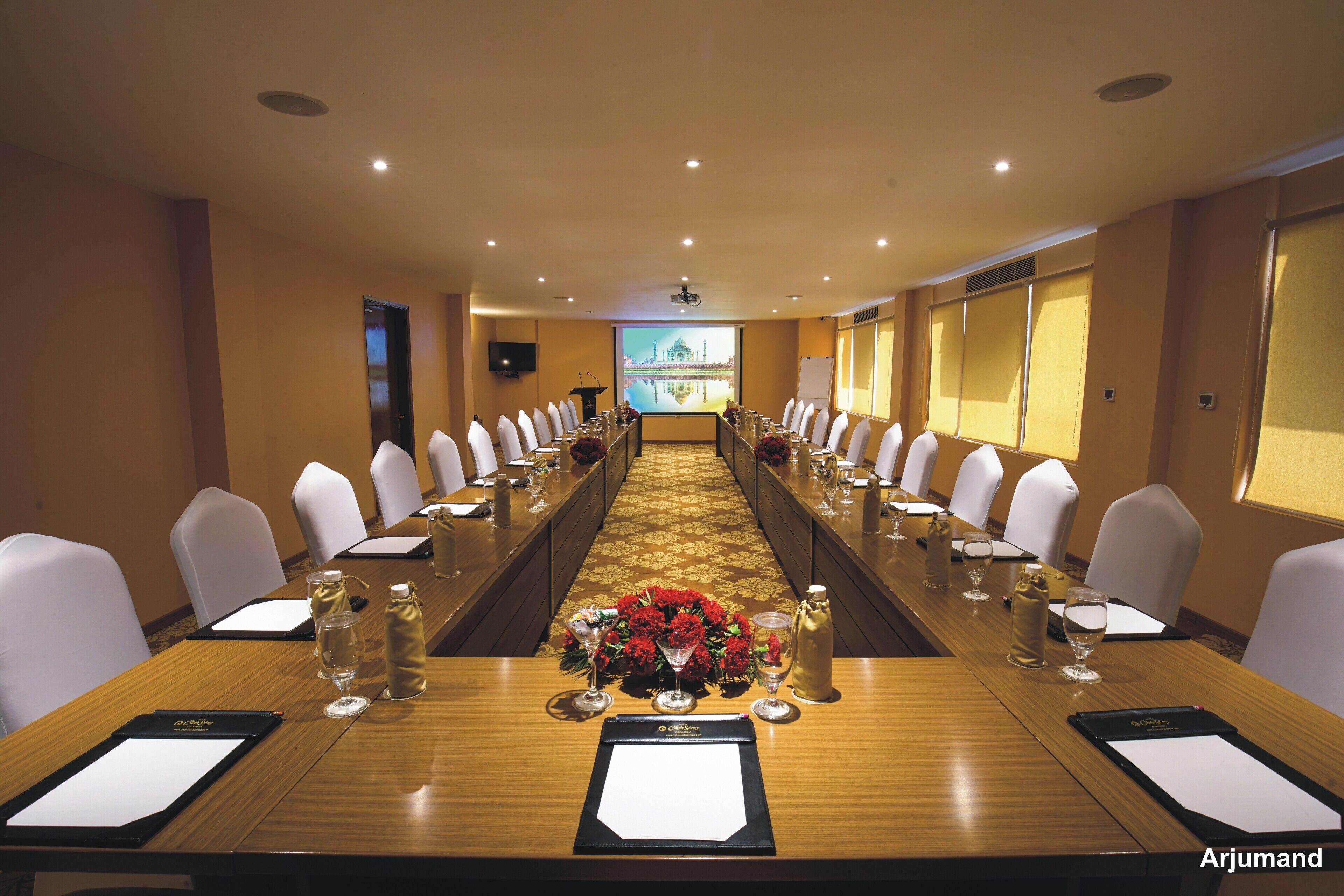 Una sala de conferencias con una larga mesa, sillas blancas y una pantalla de proyector al frente.