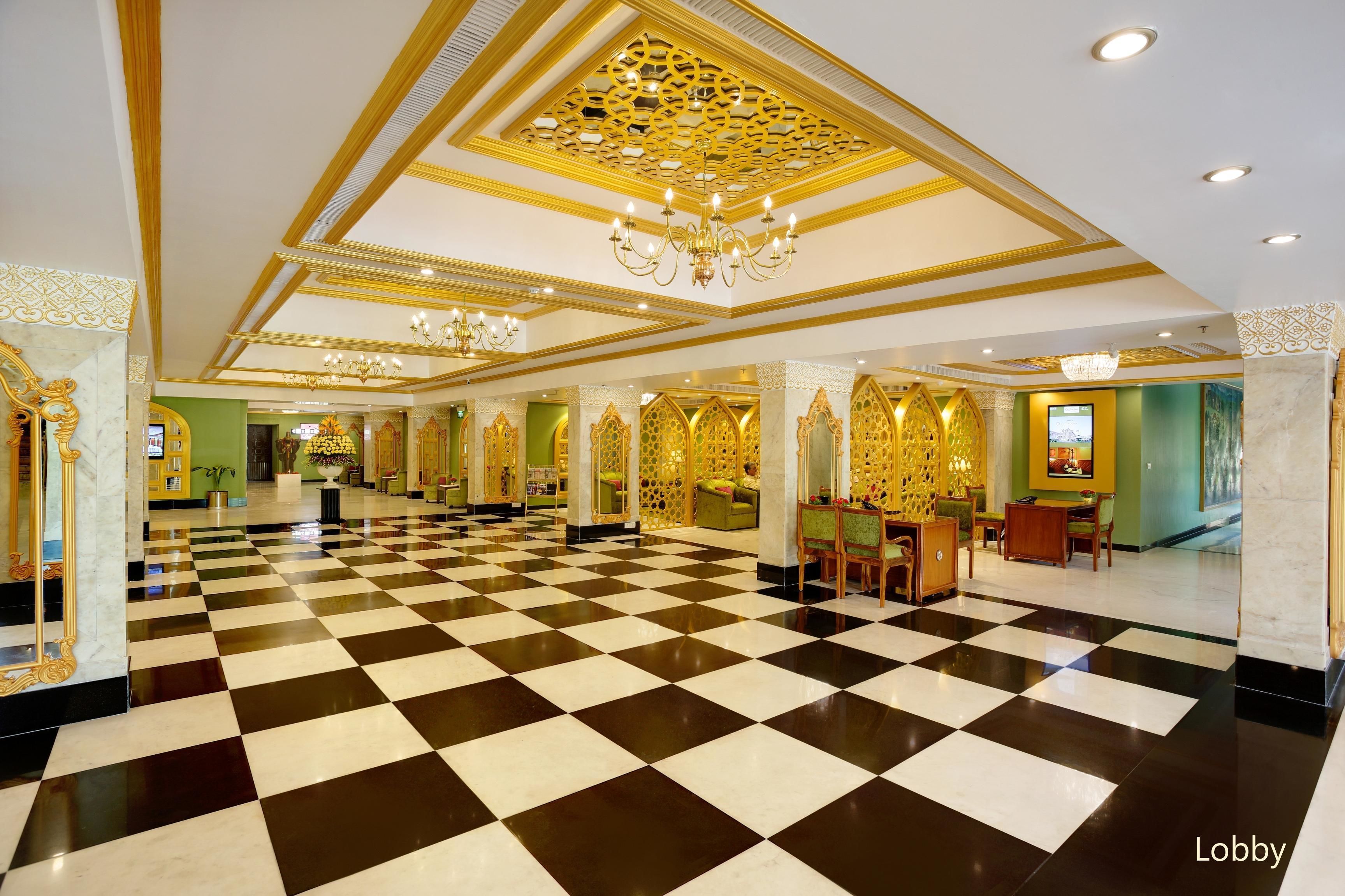 A lobby with a checkered floor, ornate gold detailing, chandeliers, and seating areas.