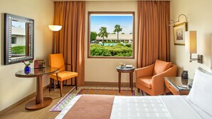 A hotel room with a bed, two chairs, a small table, a lamp, a window with a view of a pool and palm trees, and a mirror on the wall. - Trident, Agra (Agra)