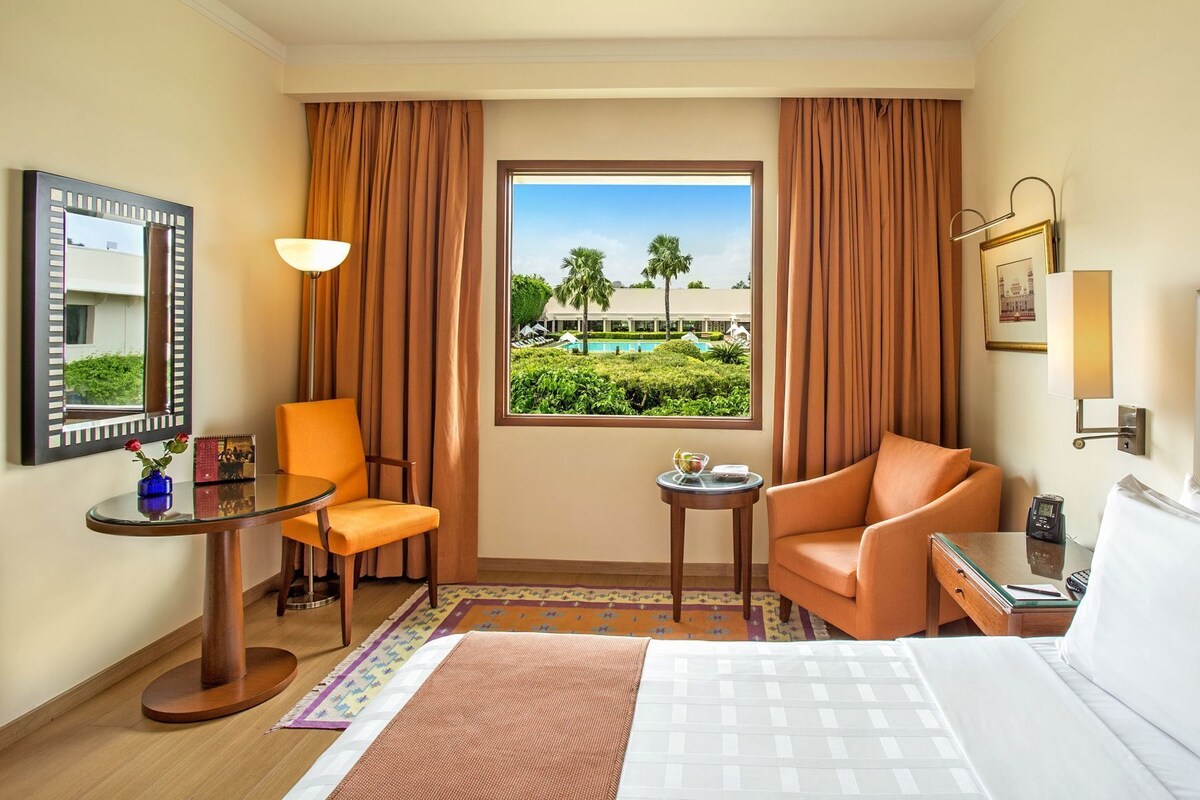 A hotel room with a bed, two armchairs, a small table, a lamp, a mirror, and a window with a view of greenery and a building.