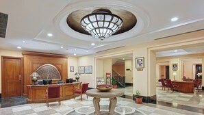A lobby with a reception desk, a round table with a bowl, a staircase, and a dining area with chairs and a table. - Cama Hotel (Ahmedabad)