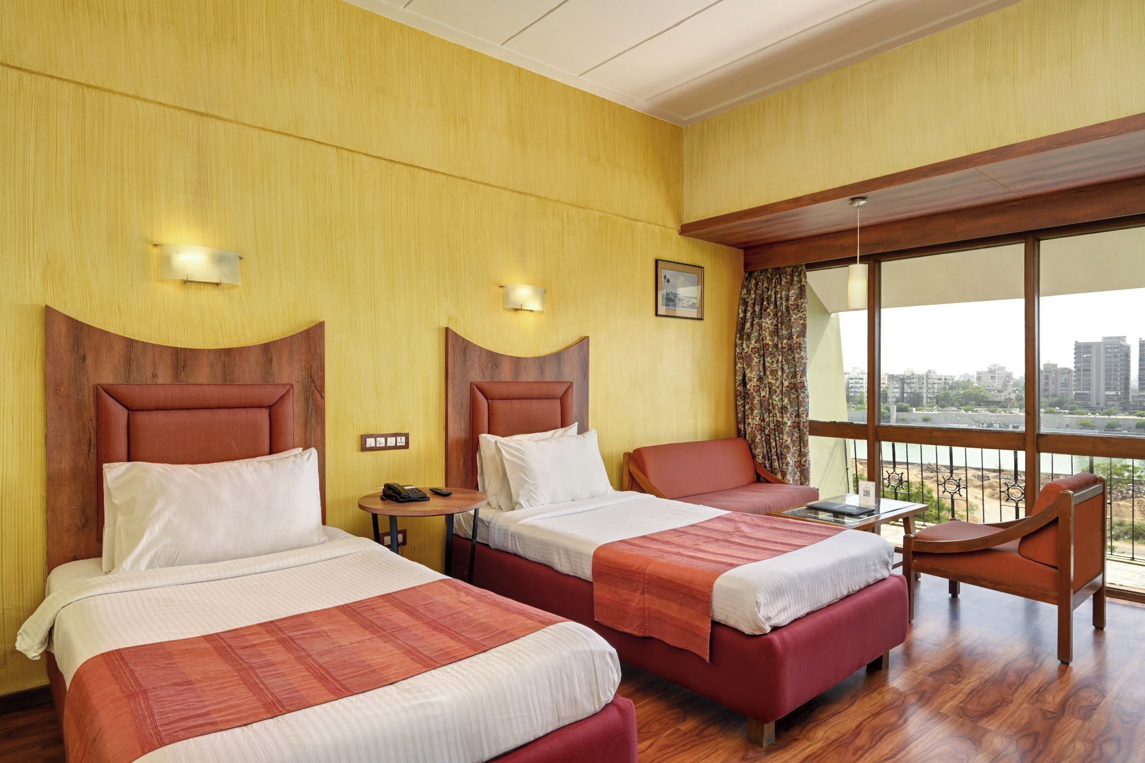 A hotel room with two beds, a small table, a chair, and a view of the city through large windows.