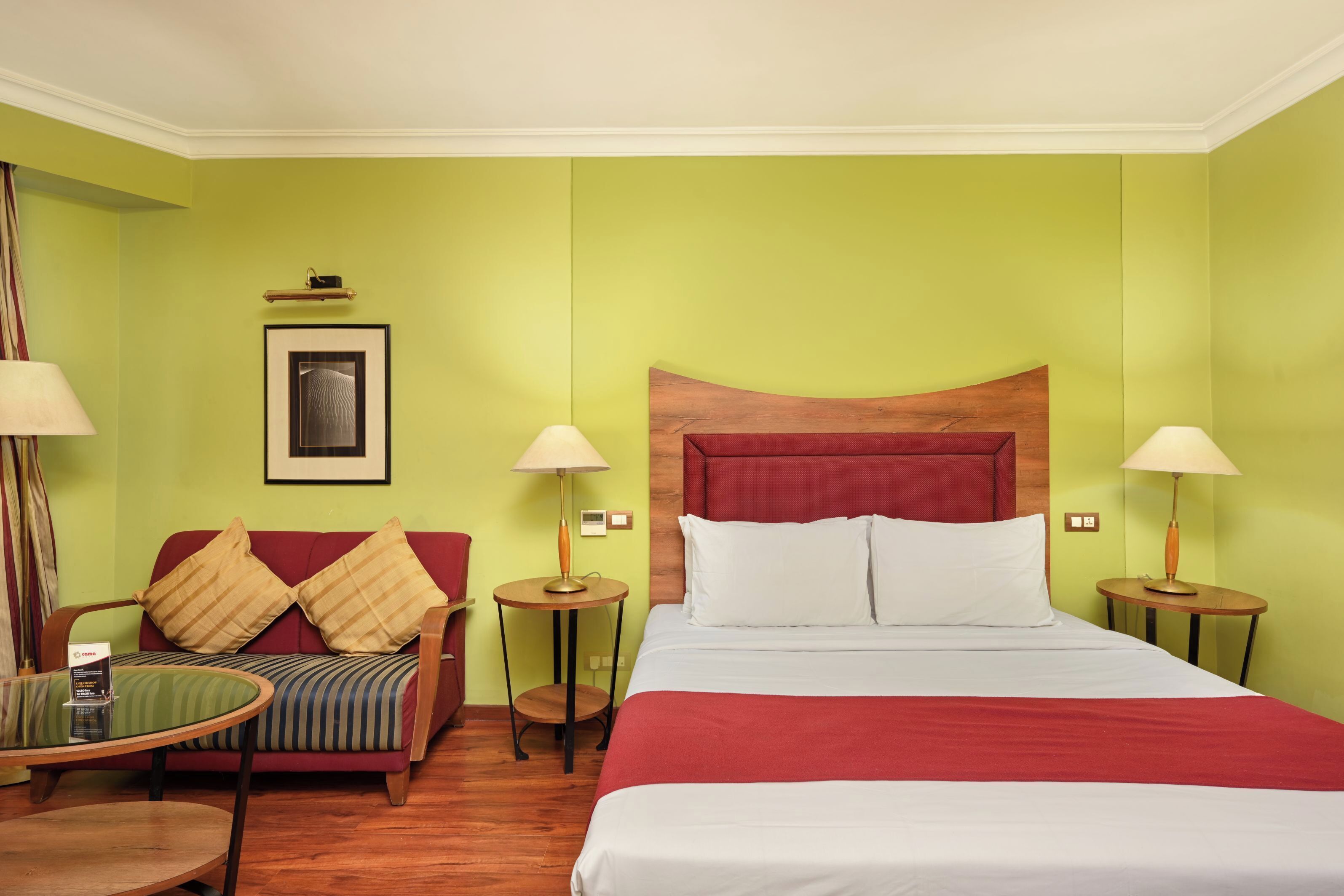 A hotel room with a bed, a sofa, two side tables with lamps, a framed picture on the wall, and a small table with a menu.