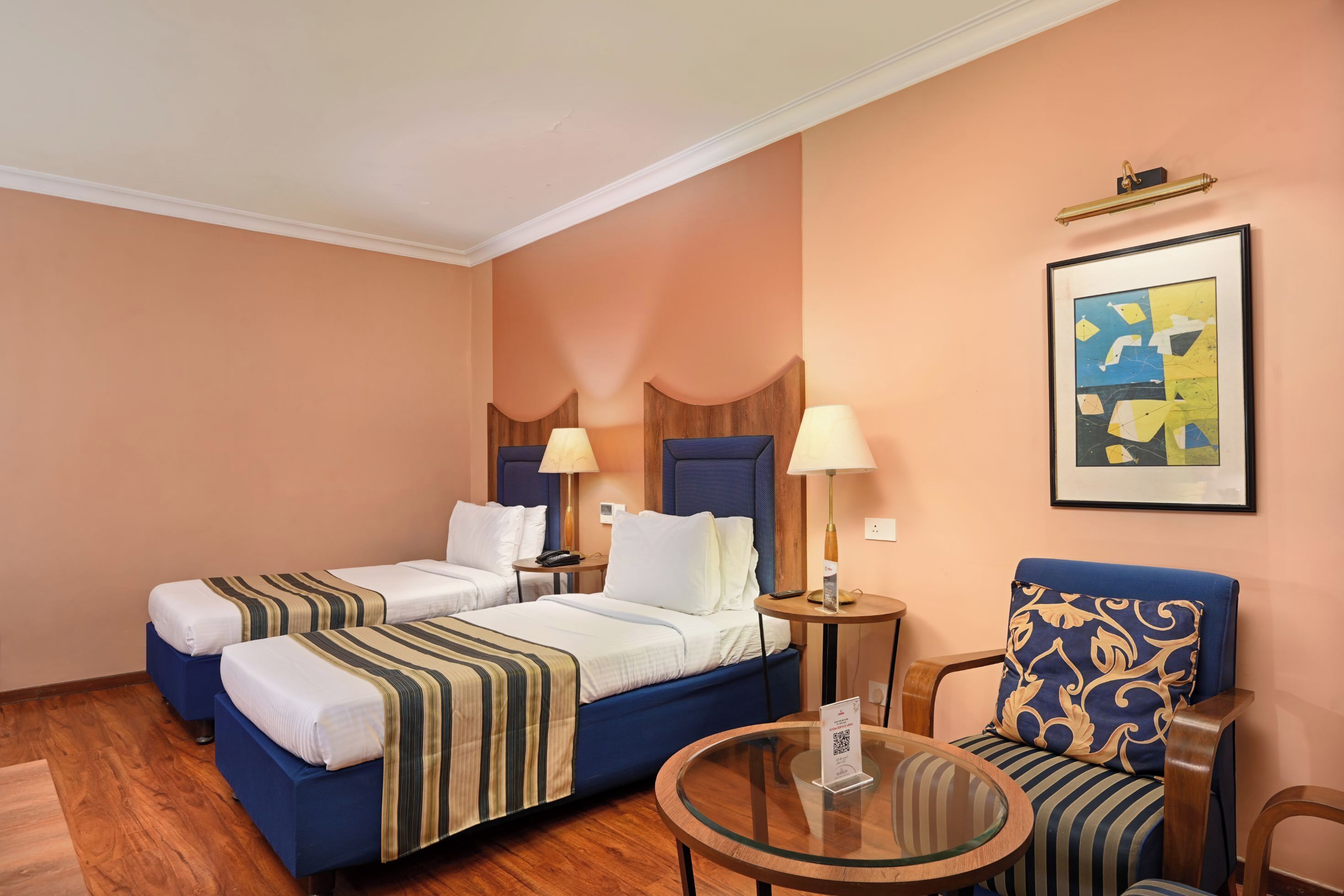 A hotel room with two beds, a small round table, a chair, and a framed abstract painting on the wall.