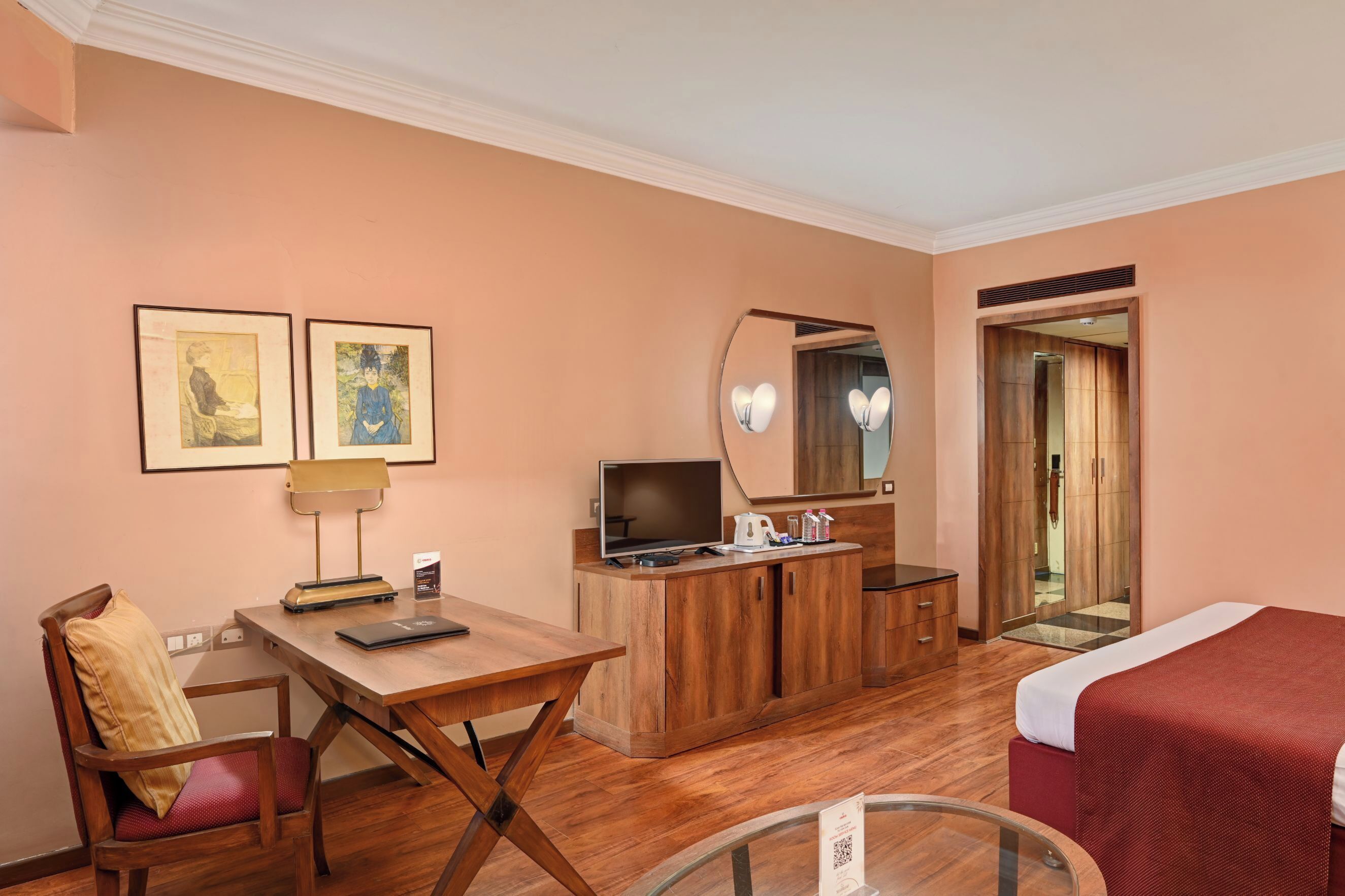 Una habitación de hotel con un escritorio de madera, una cama con cubrecama rojo, un televisor, un espejo y dos obras de arte enmarcadas en la pared.
