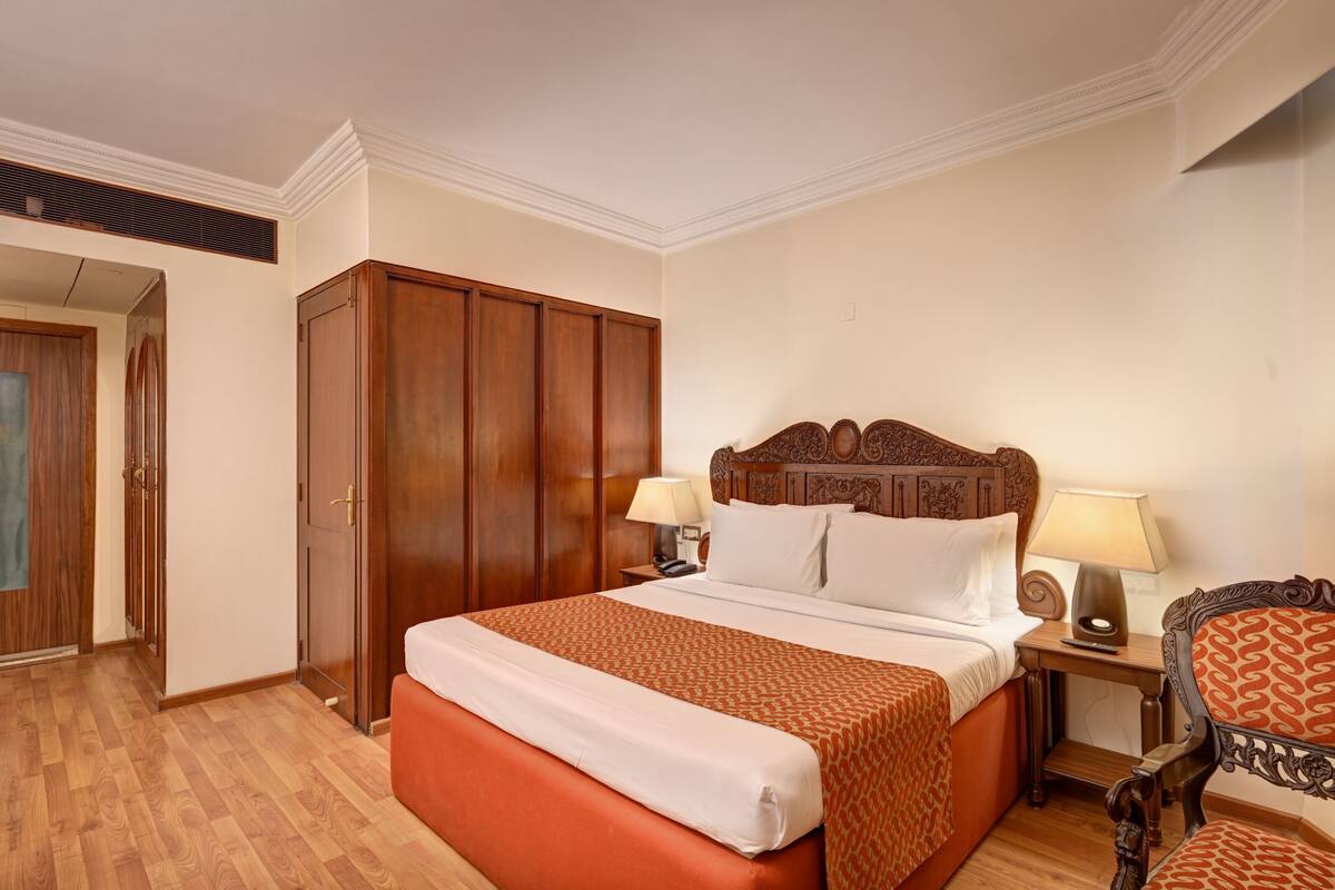 Habitación de hotel con una cama grande, un armario de madera, dos mesitas de noche con lámparas y una silla estampada.