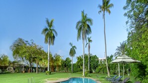 A swimming pool surrounded by grass, palm trees, and lounge chairs. - Cama Hotel (Ahmedabad)