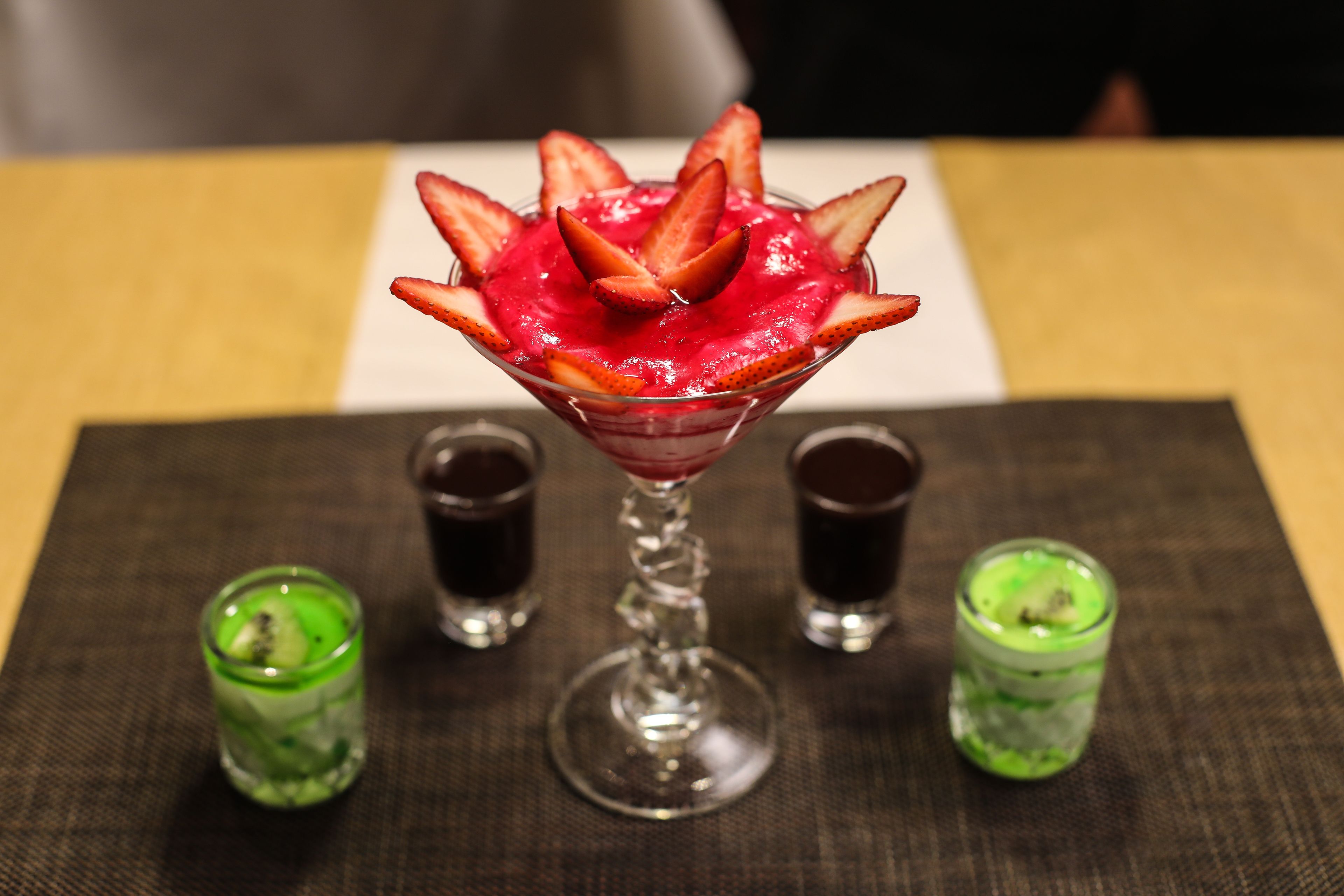 Een dessert in een glazen schaal, met aardbeien bovenop, omringd door shotglazen op een tafel.