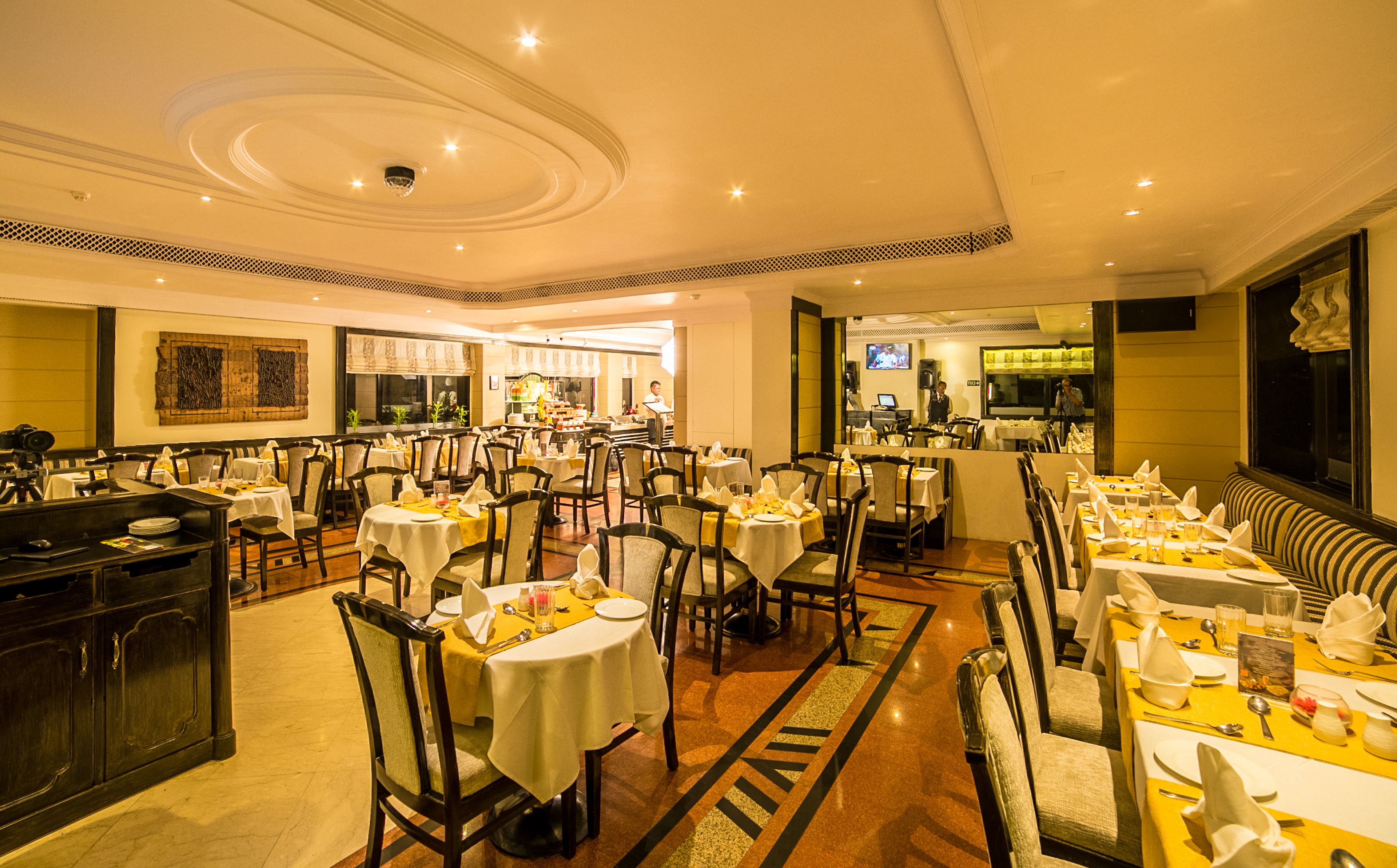 A spacious restaurant with multiple dining tables and chairs, a kitchen area in the background, and a well-lit interior.
