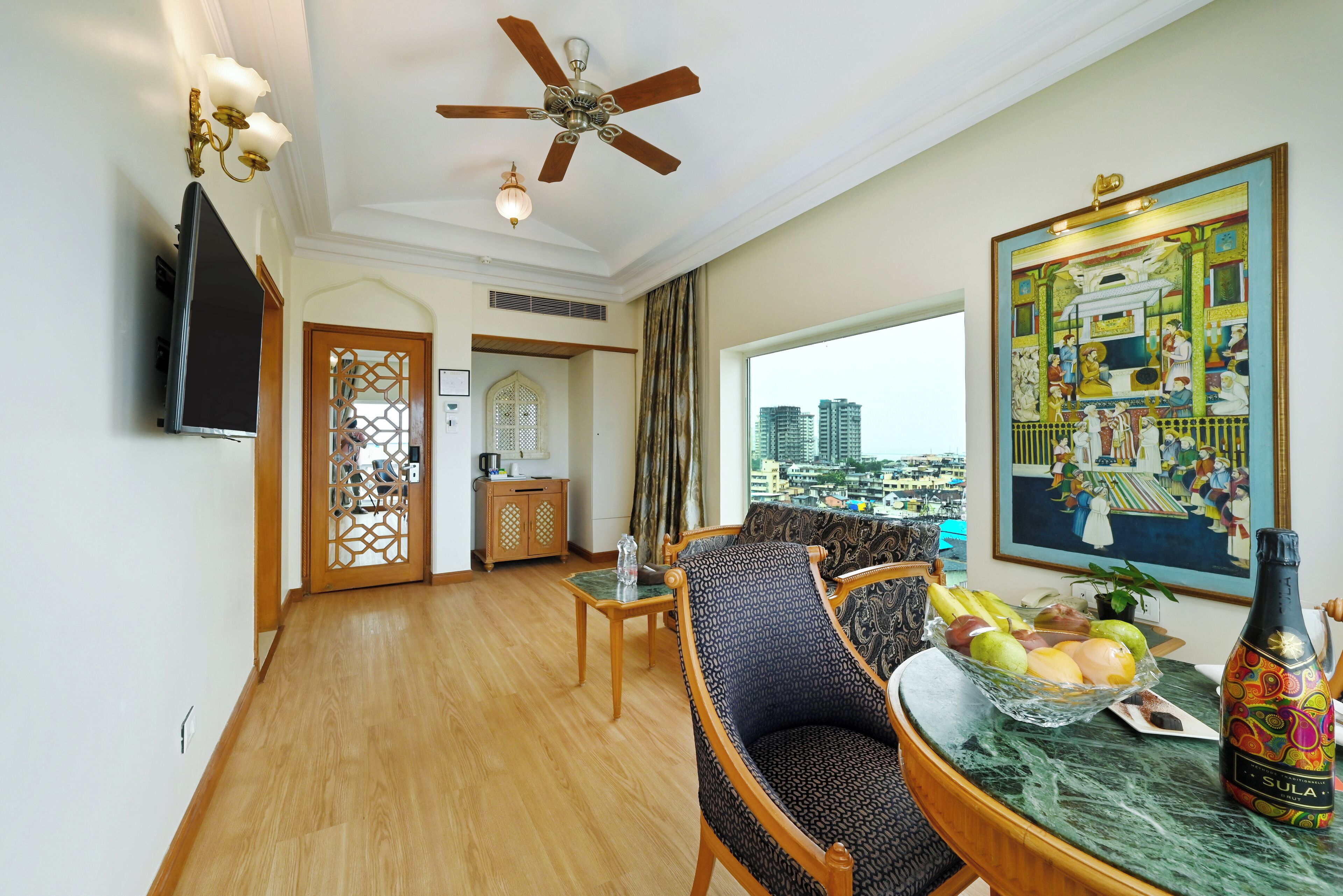 A spacious living room with a ceiling fan, a television, a dining table with fruit and a bottle, and a large window with a city view.