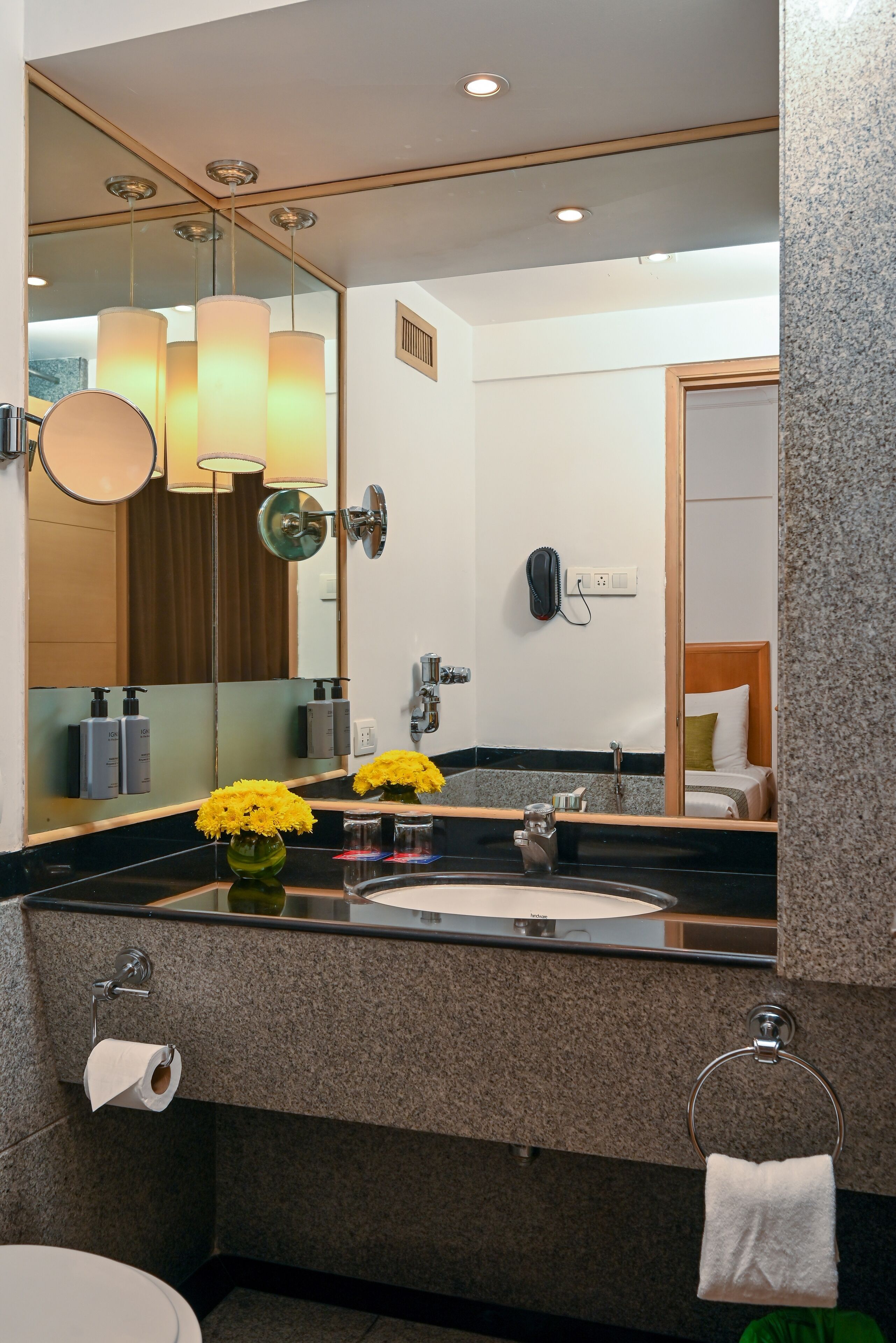 A modern bathroom with a large mirror, a sink, and a toilet. There are yellow flowers on the counter, a towel rack, and a phone on the wall.