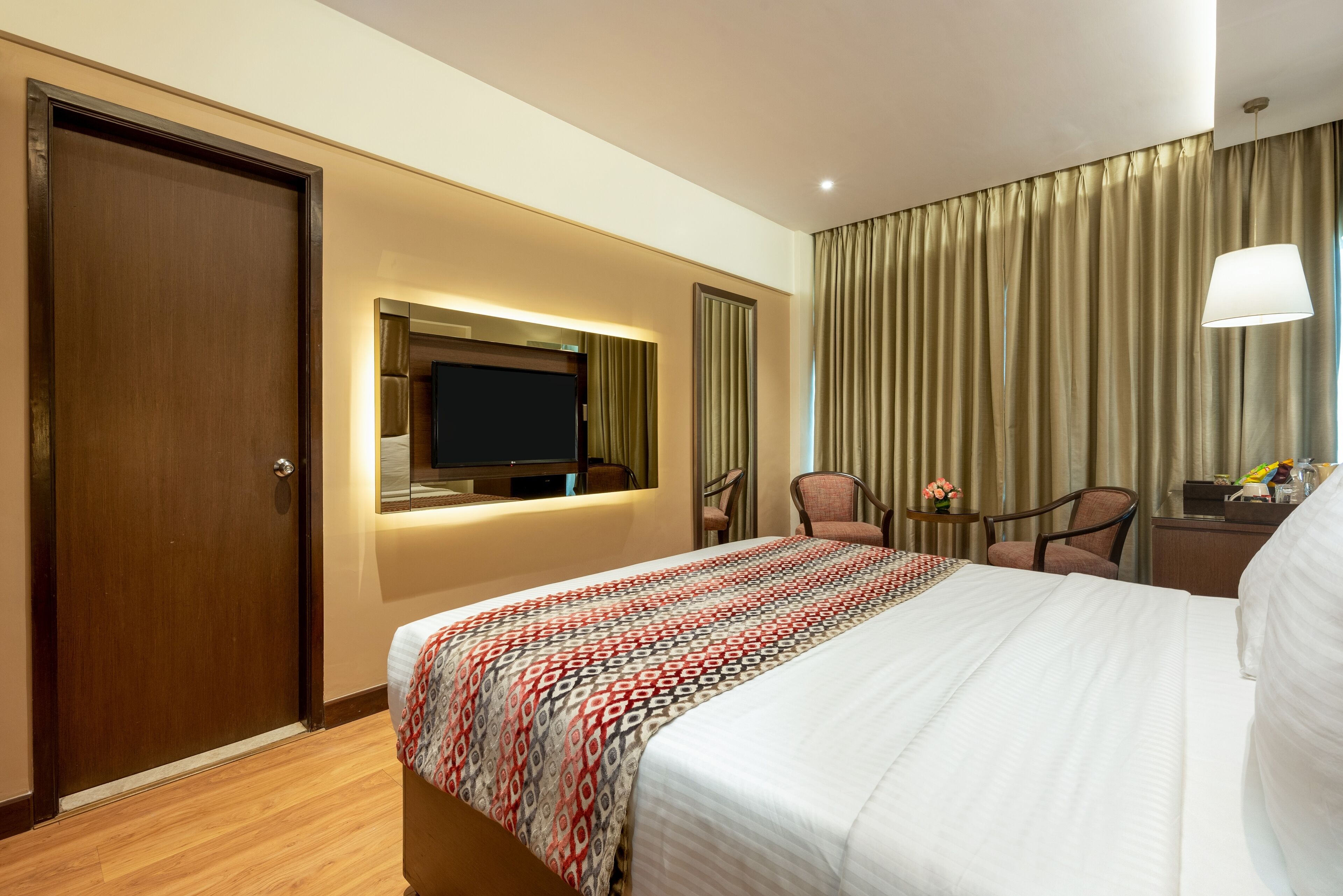 Habitación de hotel con una cama grande, un televisor montado en la pared, un escritorio con lámpara y vistas al exterior a través de cortinas.