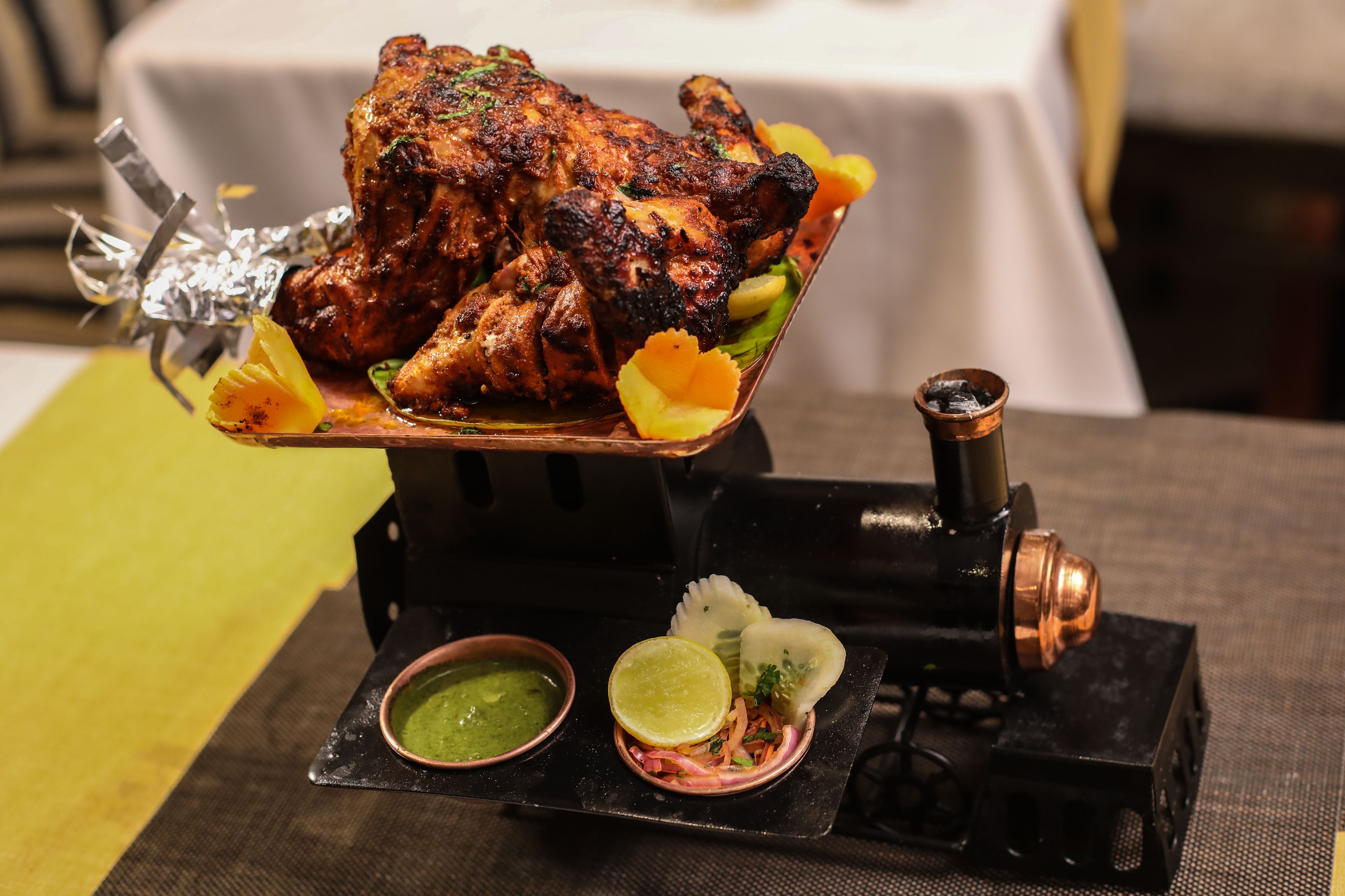 Un poulet rôti, présenté sur un présentoir en métal, accompagné d’une sauce verte et de tranches de citron vert.
