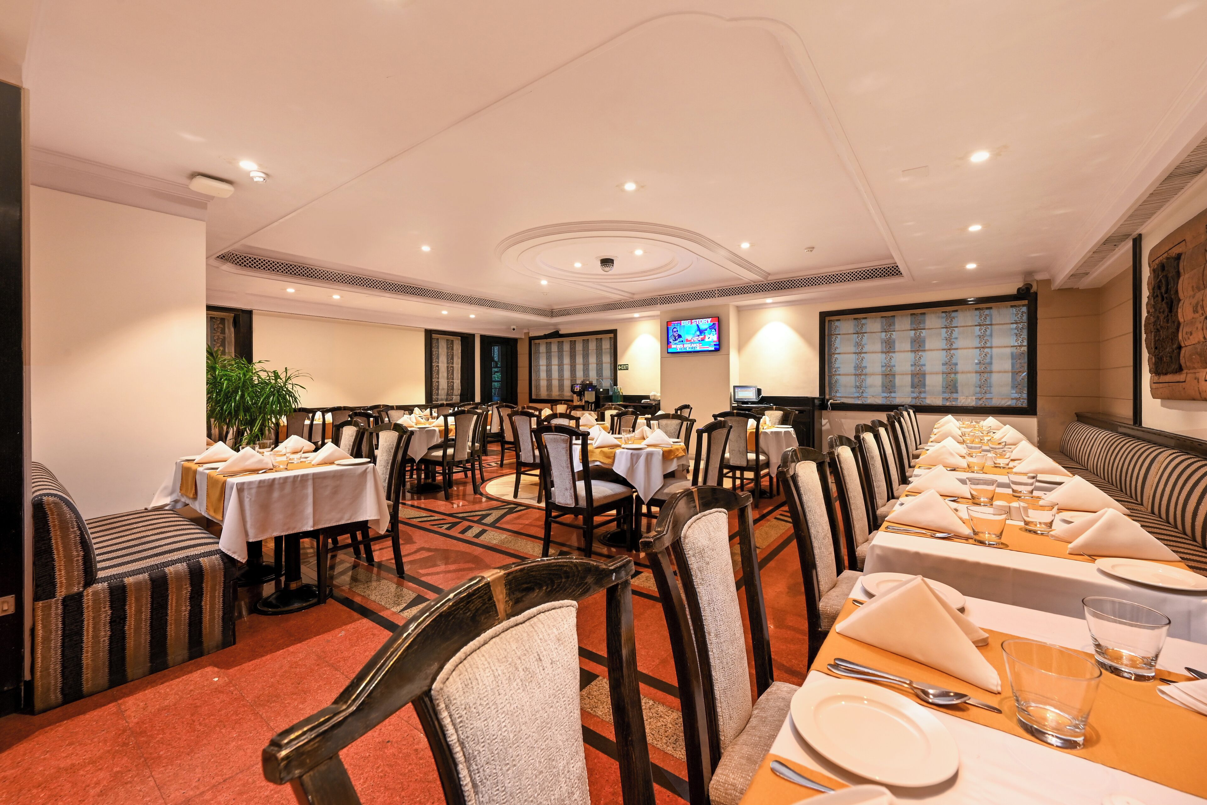 Een restaurantgedeelte van een hotel met meerdere gedekte tafels voor gasten, stoelen en een televisiescherm aan de muur.