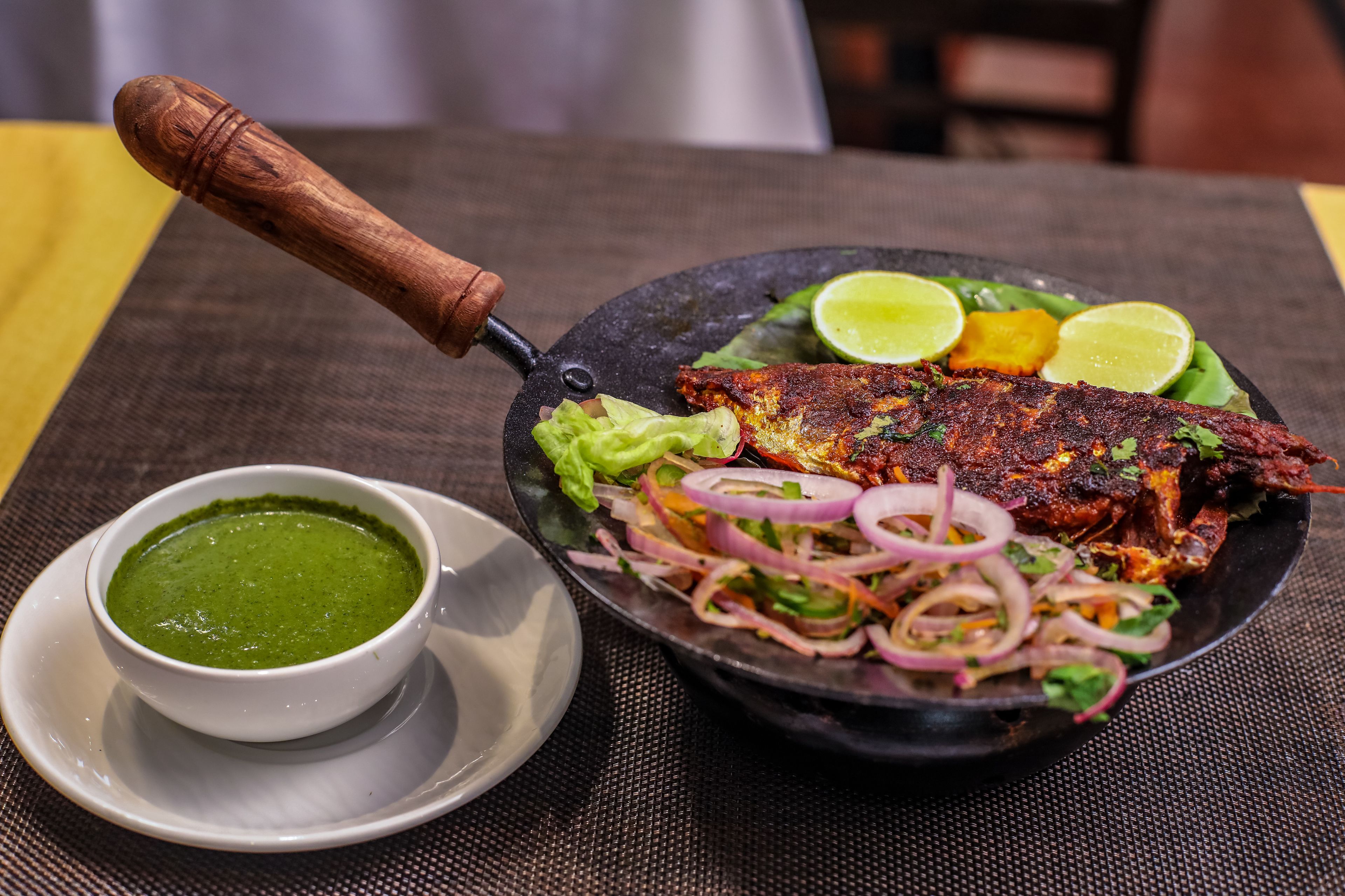Un plato con pescado a la parrilla, ensalada y un tazón de salsa verde sobre una mesa.