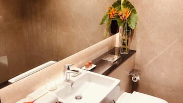 A modern bathroom with a large mirror, white sink, and toilet. There are towels and a vase with flowers on the counter.