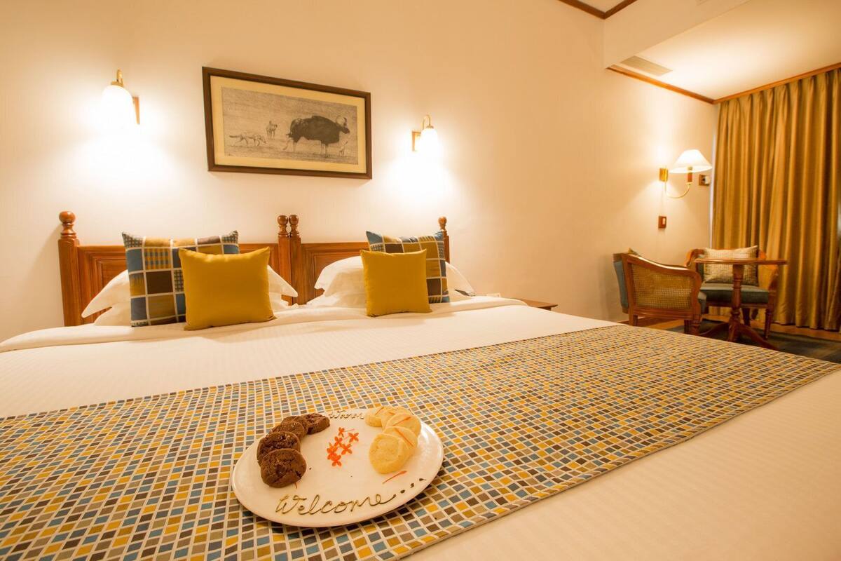 Una habitación de hotel con una cama, un cuadro en la pared, una silla, y un plato con galletas y un pastel sobre la cama.