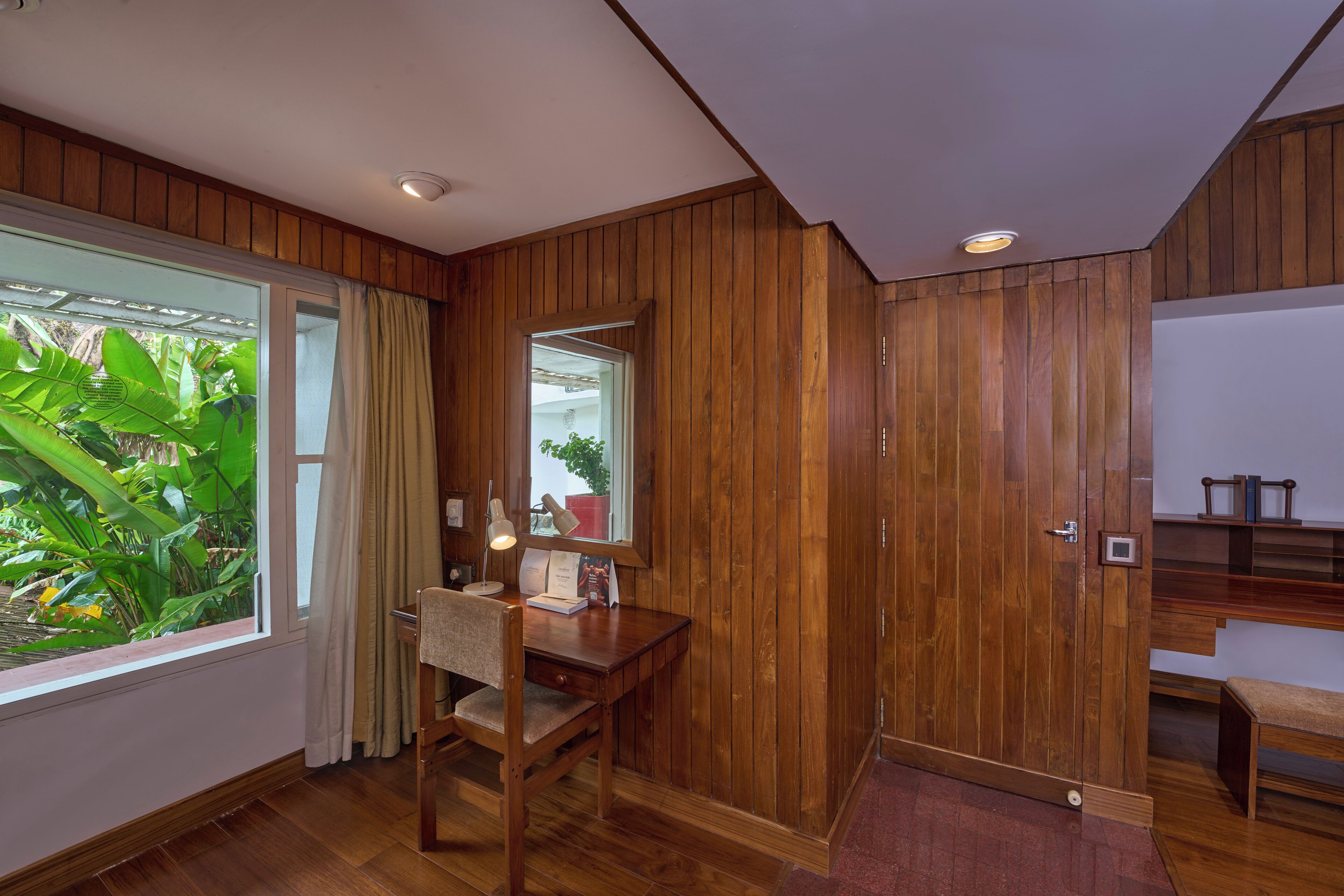A room with wooden paneling, a desk with a chair, a window with curtains, and a view of greenery outside.