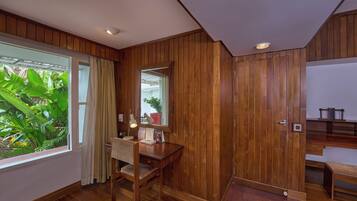A room with wooden paneling, a desk with a chair, a window with curtains, and a view of greenery outside.