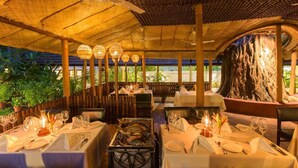 Un restaurante al aire libre con mesas preparadas para comer, un tronco de árbol grande al fondo y vista al mar.