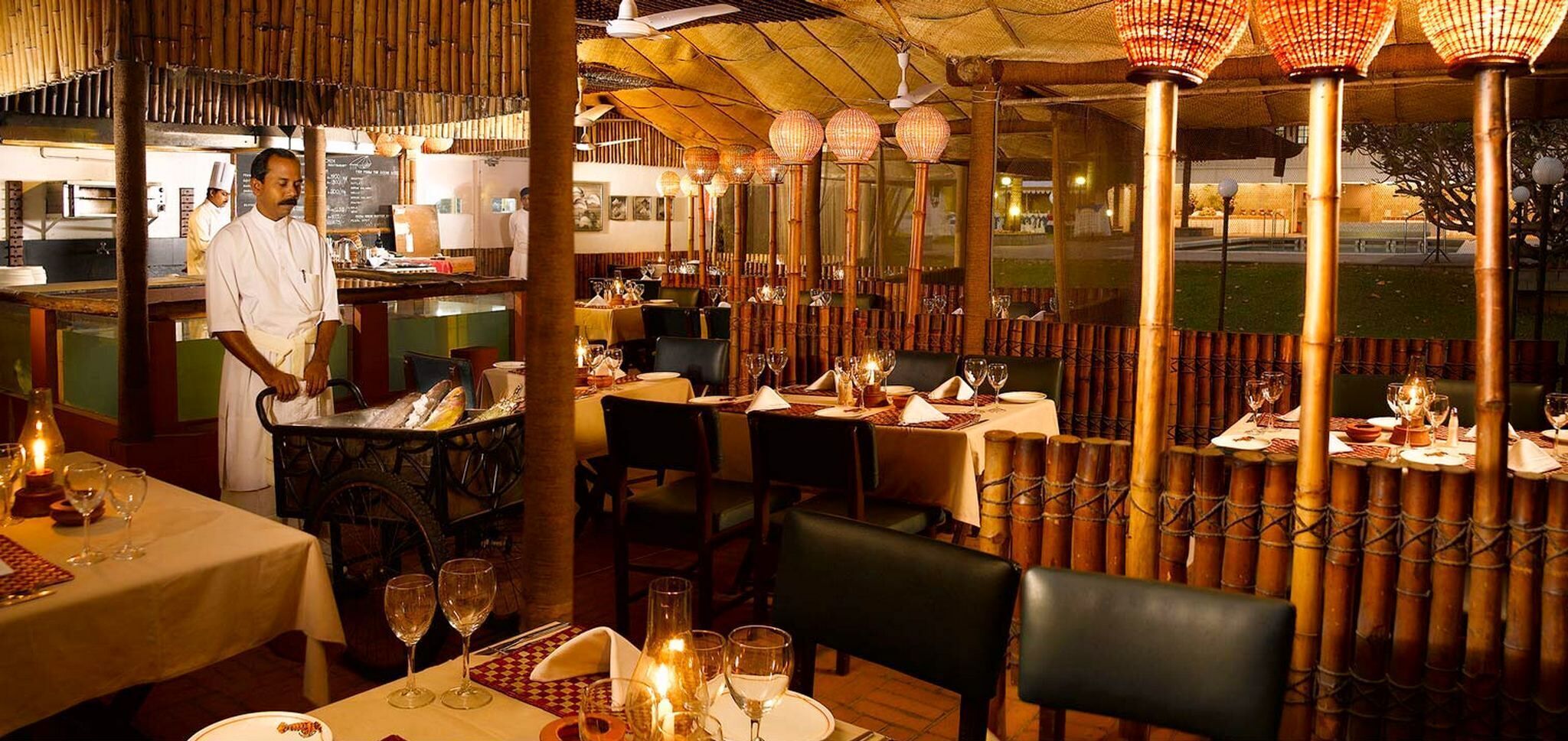 Un restaurante interior con separadores de bambú, mesas dispuestas para comer y un cocinero preparando comida.