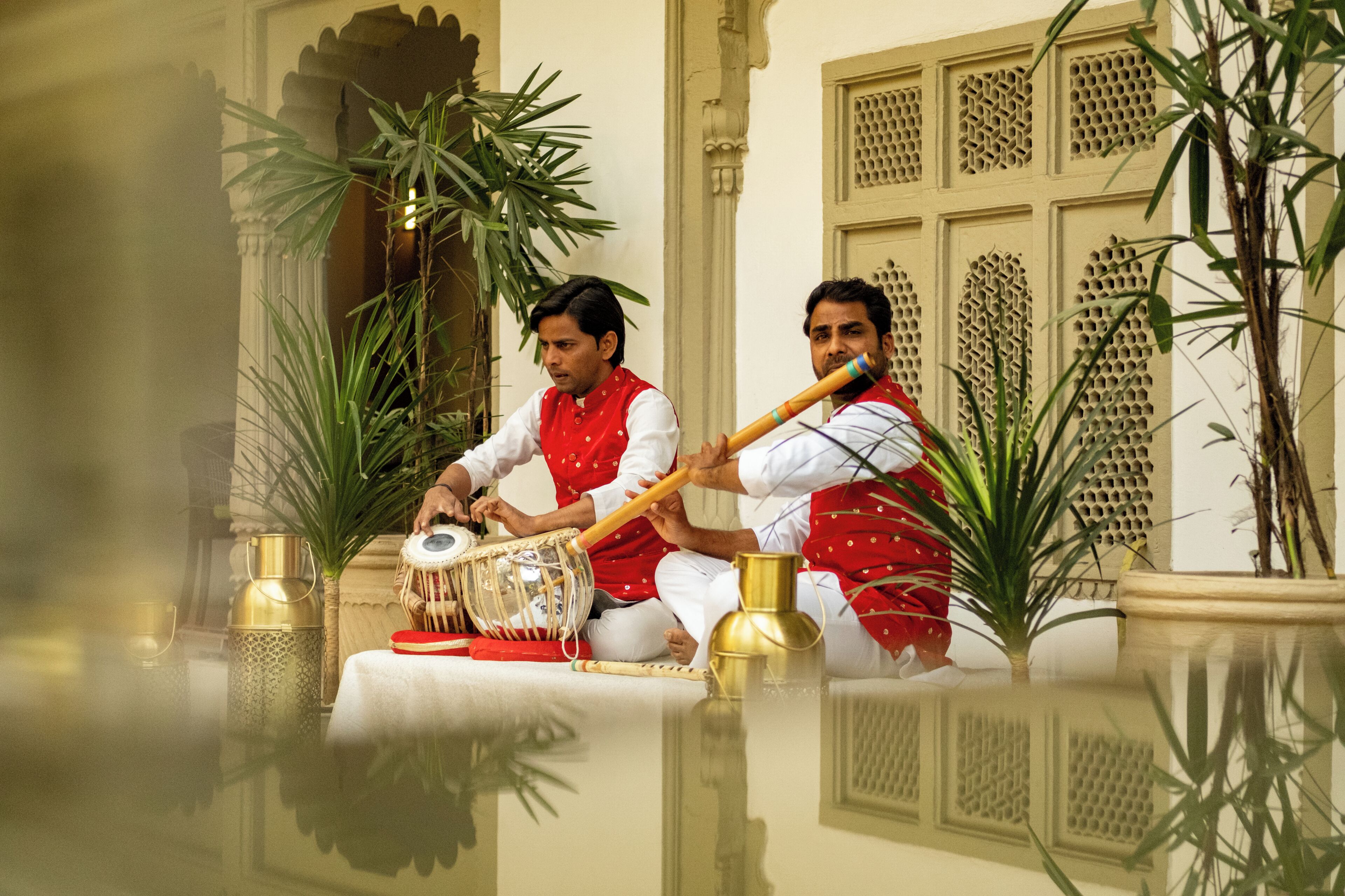Two individuals seated on a reflective surface, playing traditional musical instruments, with architectural elements and potted plants in the background.