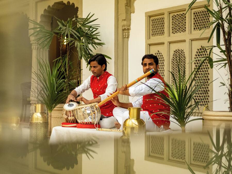 Dos personas sentadas sobre una superficie reflectante, tocando instrumentos musicales tradicionales, con elementos arquitectónicos y plantas en macetas al fondo.