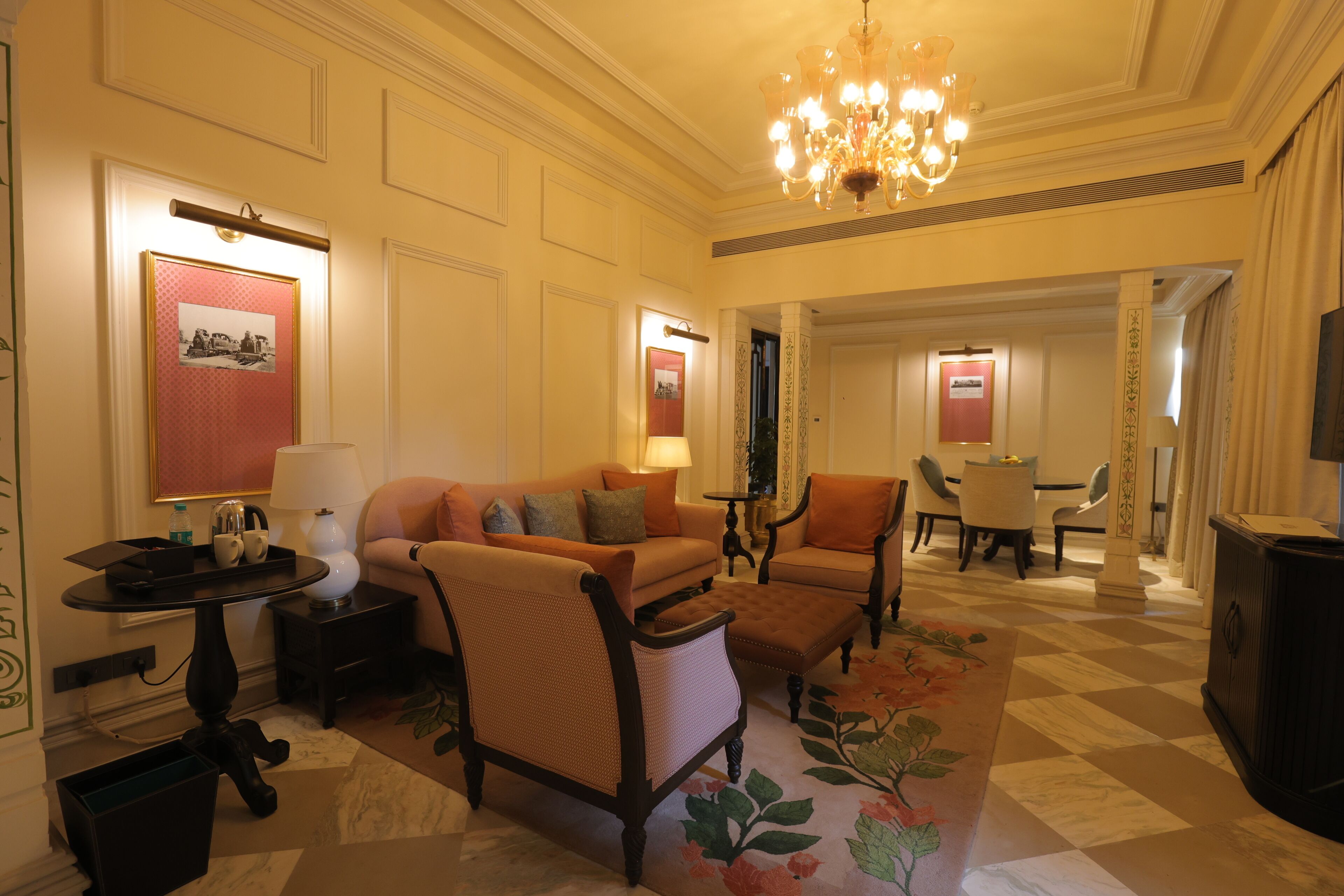 A hotel lobby with a sofa, armchairs, a table, a chandelier, and a dining area in the background.