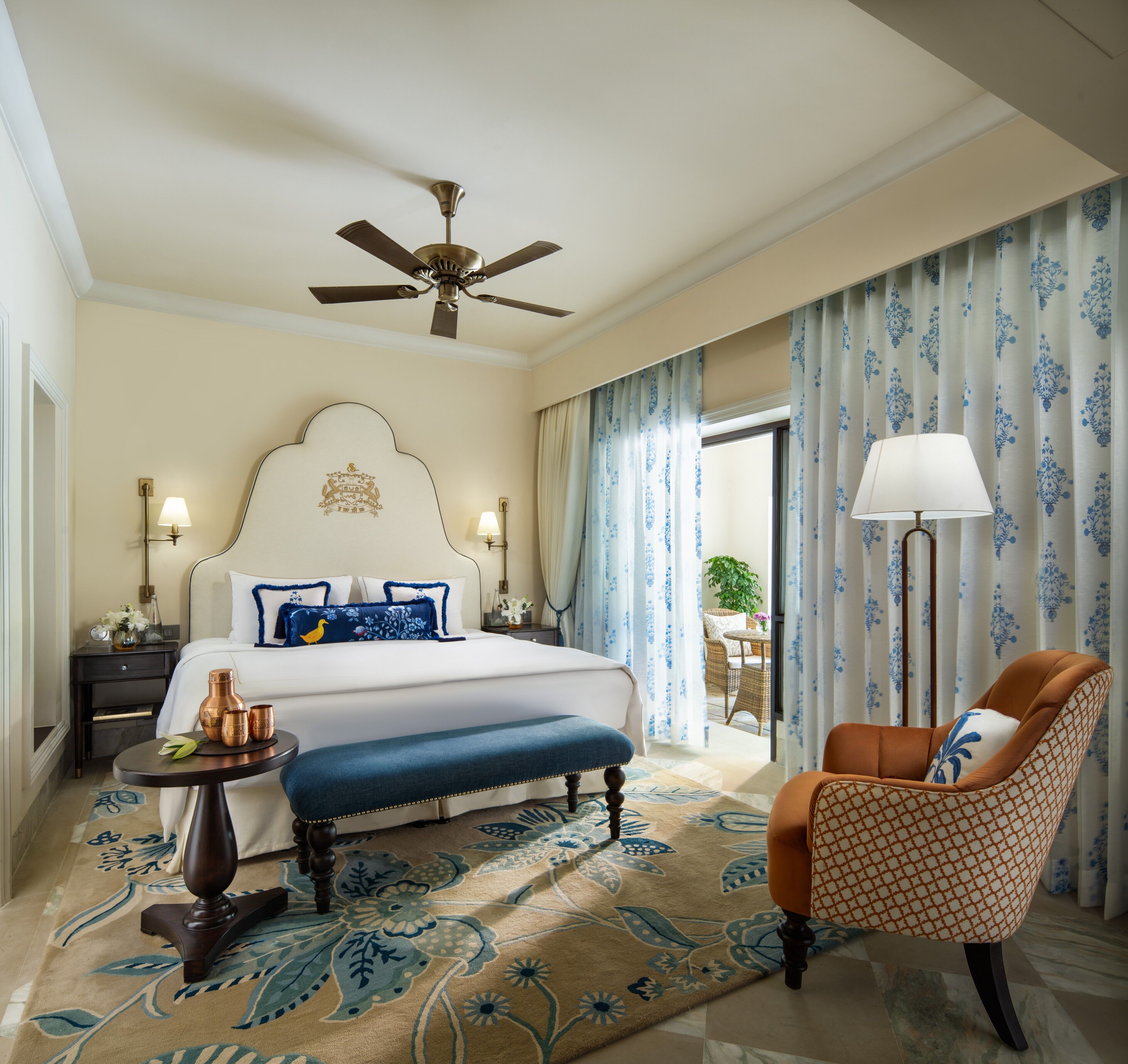 Habitación de hotel con una cama grande, ventilador de techo, zona de estar con una silla, una mesa y vistas al exterior.