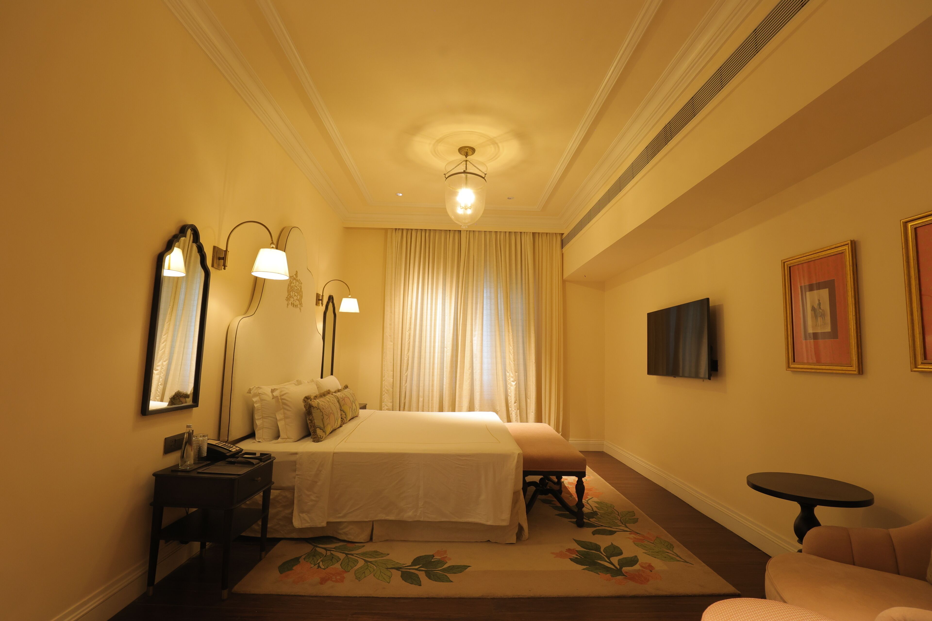A hotel room with a bed, bedside tables, a desk, a chair, a small table, a television, and a large window with curtains.